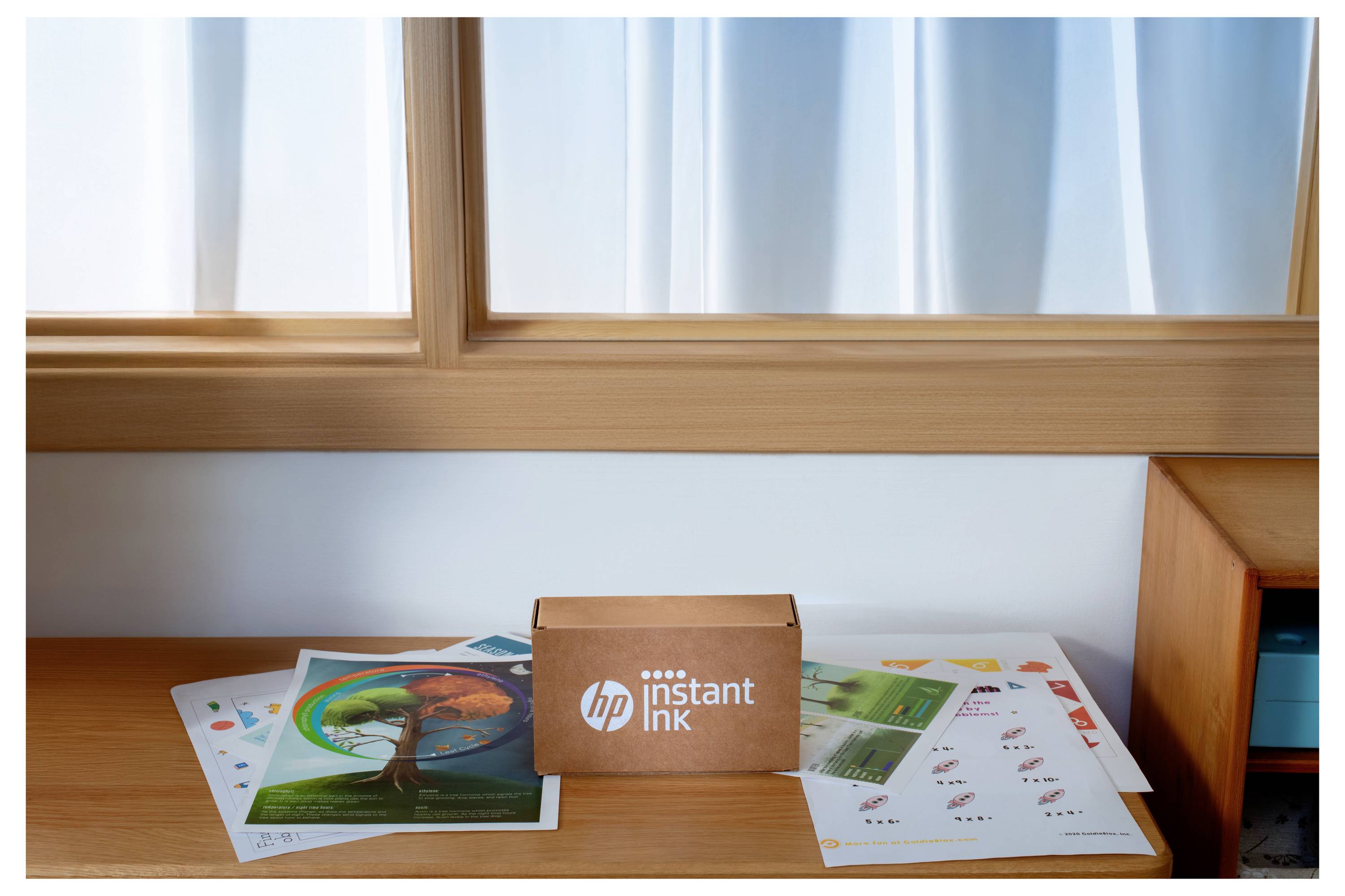 'HP Instant Ink'-doos op een houten tafel, omringd door kleurrijke gedrukte documenten, naast een raam met witte gordijnen.