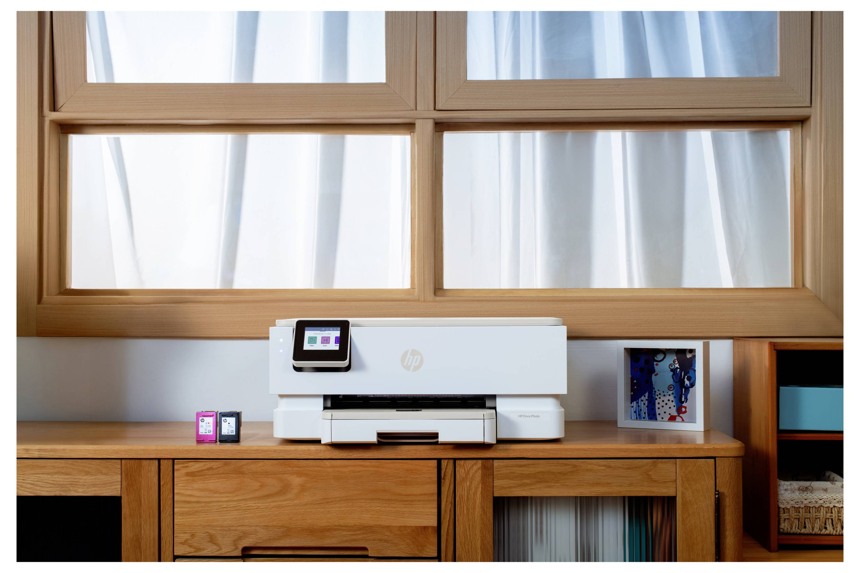 'Een printer staat op een houten bureau onder een groot raam, met twee inktpatronen ernaast en een klein ingelijst plaatje aan de rechterkant.'