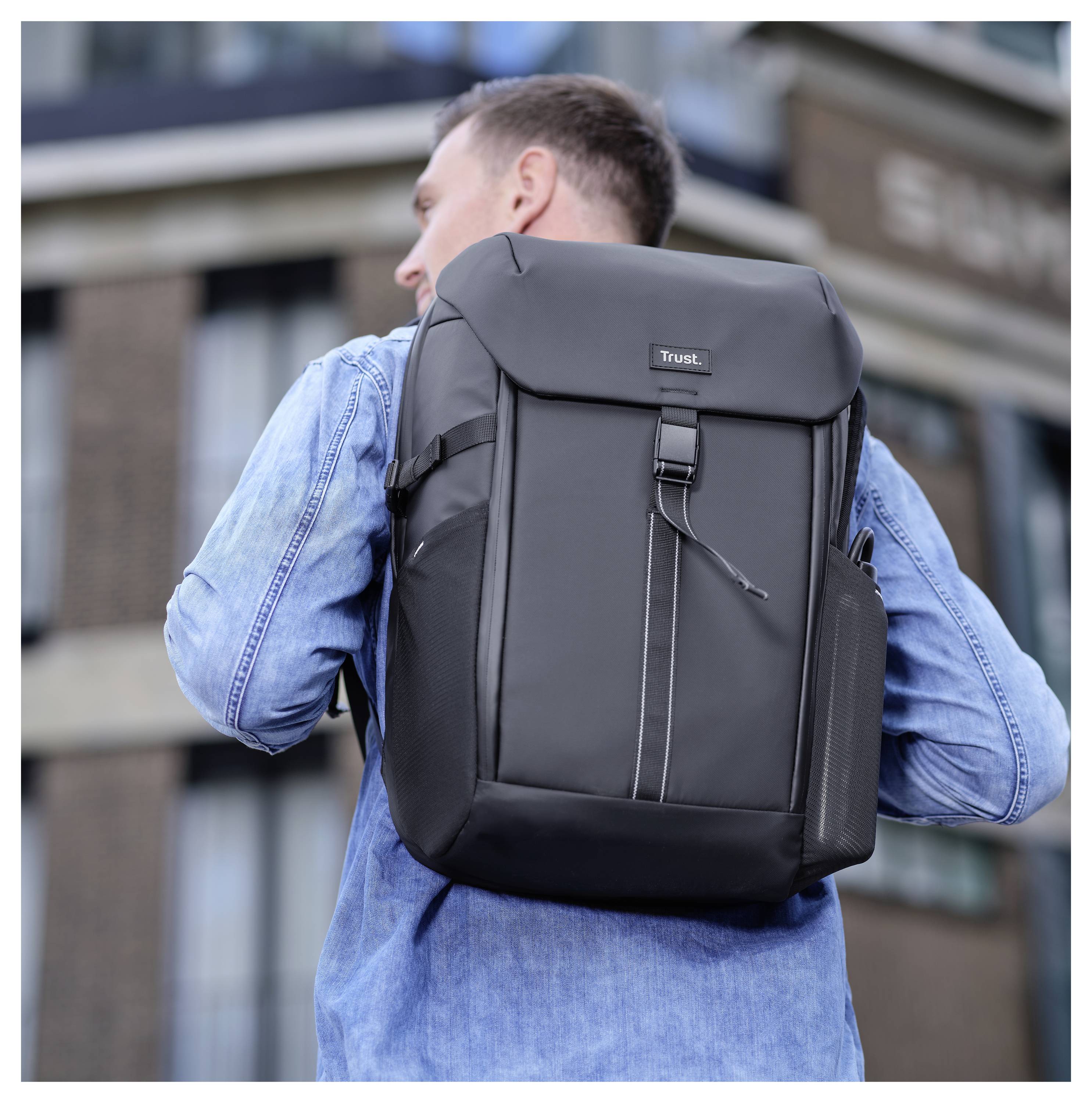 Een persoon met een spijkerjasje draagt een grote zwarte rugzak met een logo op de achterkant, staande voor een modern gebouw.