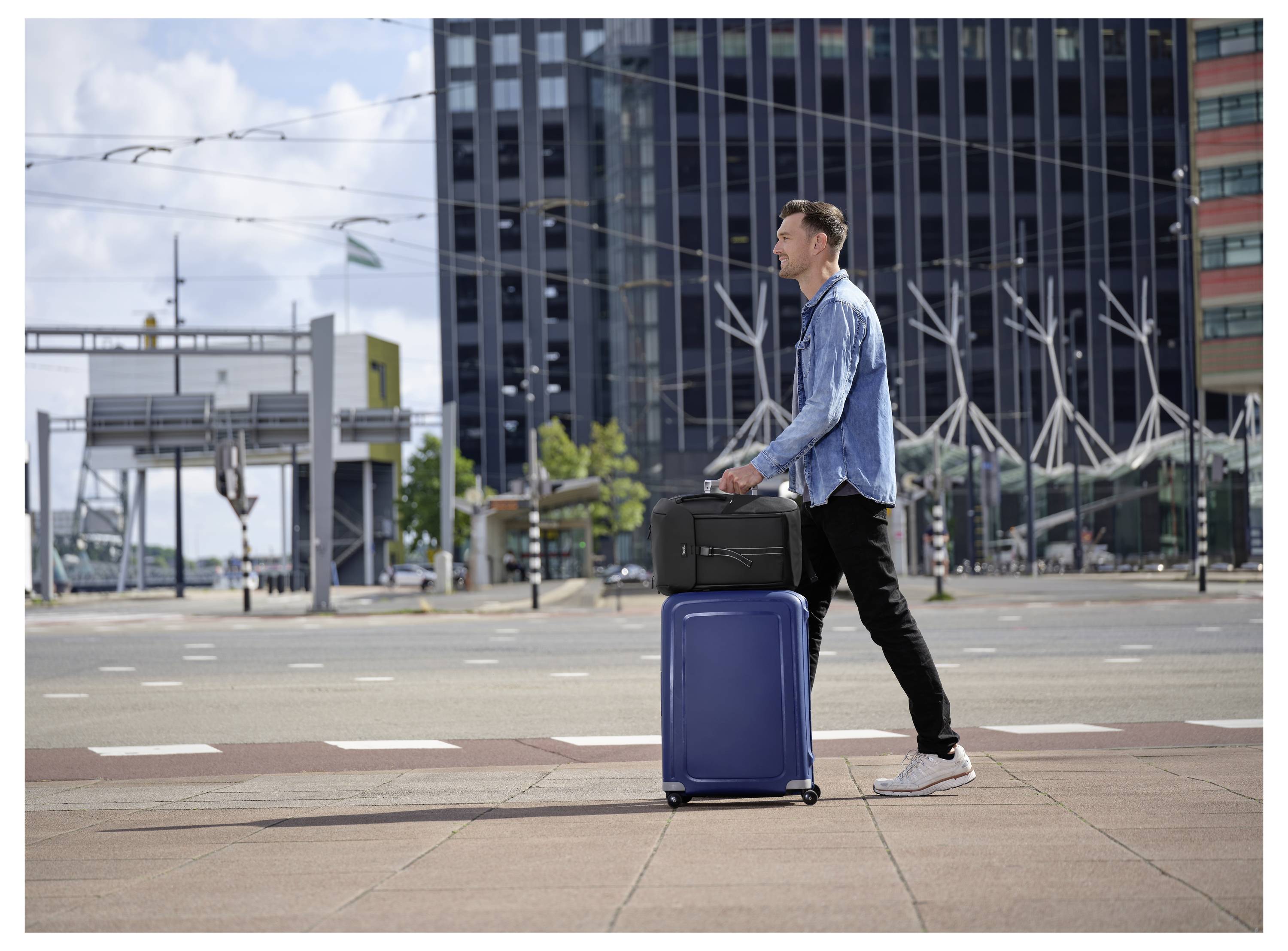 Een persoon steekt een stedelijke straat over terwijl hij twee koffers meetrekt, één blauw en één zwart, met gebouwen en een heldere lucht op de achtergrond.
