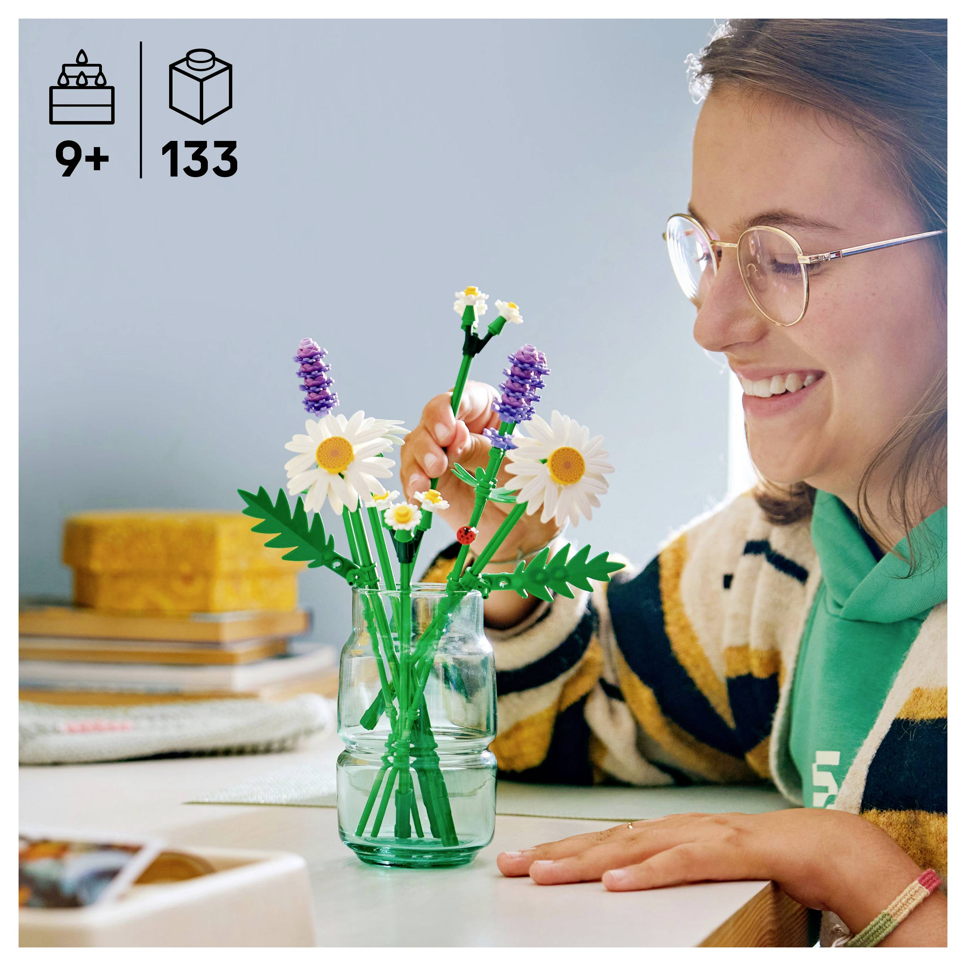 Een persoon die een LEGO bloemset met madeliefjes en lavendelbloemen in elkaar aan het zetten is, zittend aan een tafel met een vreugdevolle uitdrukking.