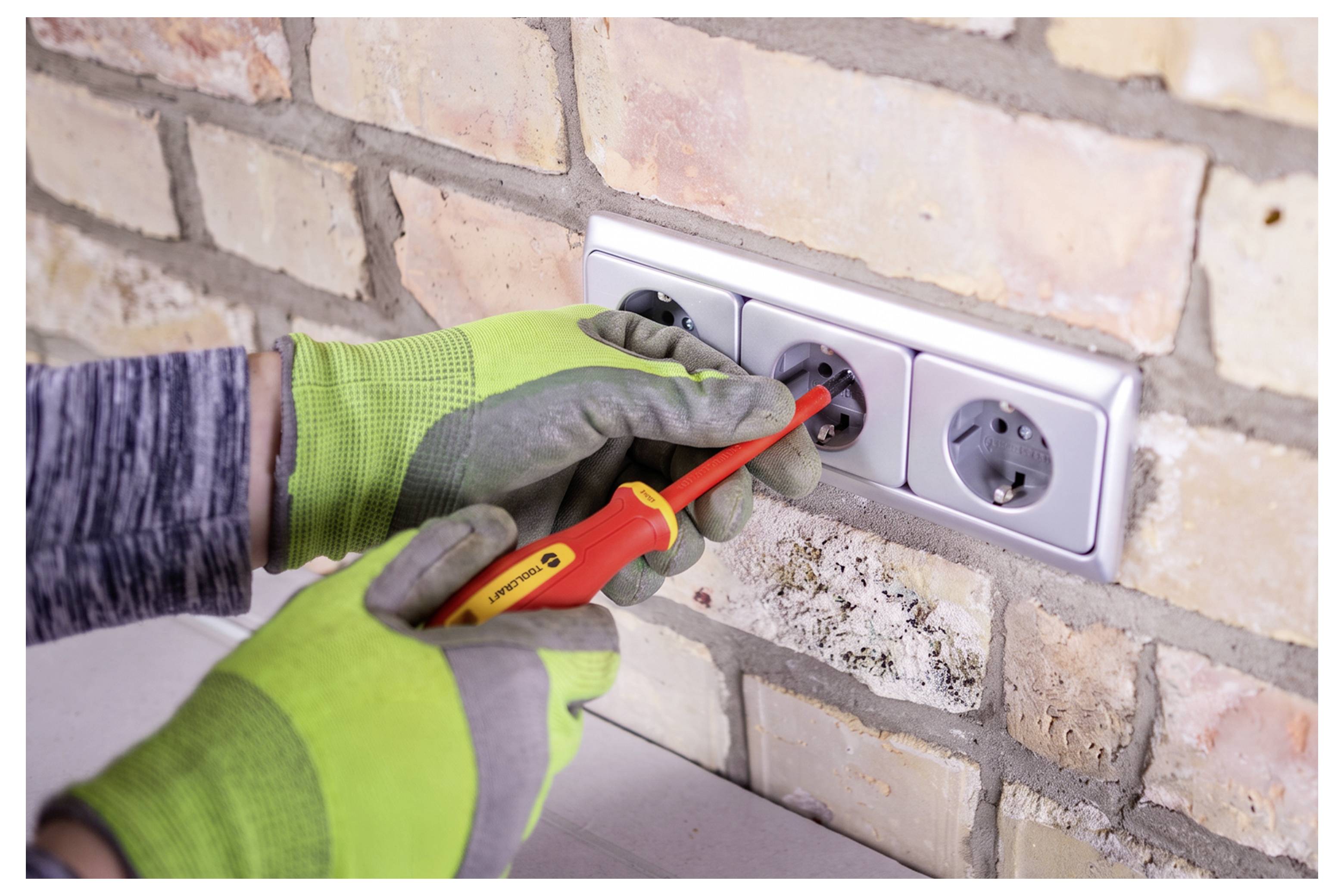 Gehandschoende werker gebruikt een schroevendraaier om een elektrisch stopcontact op een bakstenen muur te testen of te installeren.