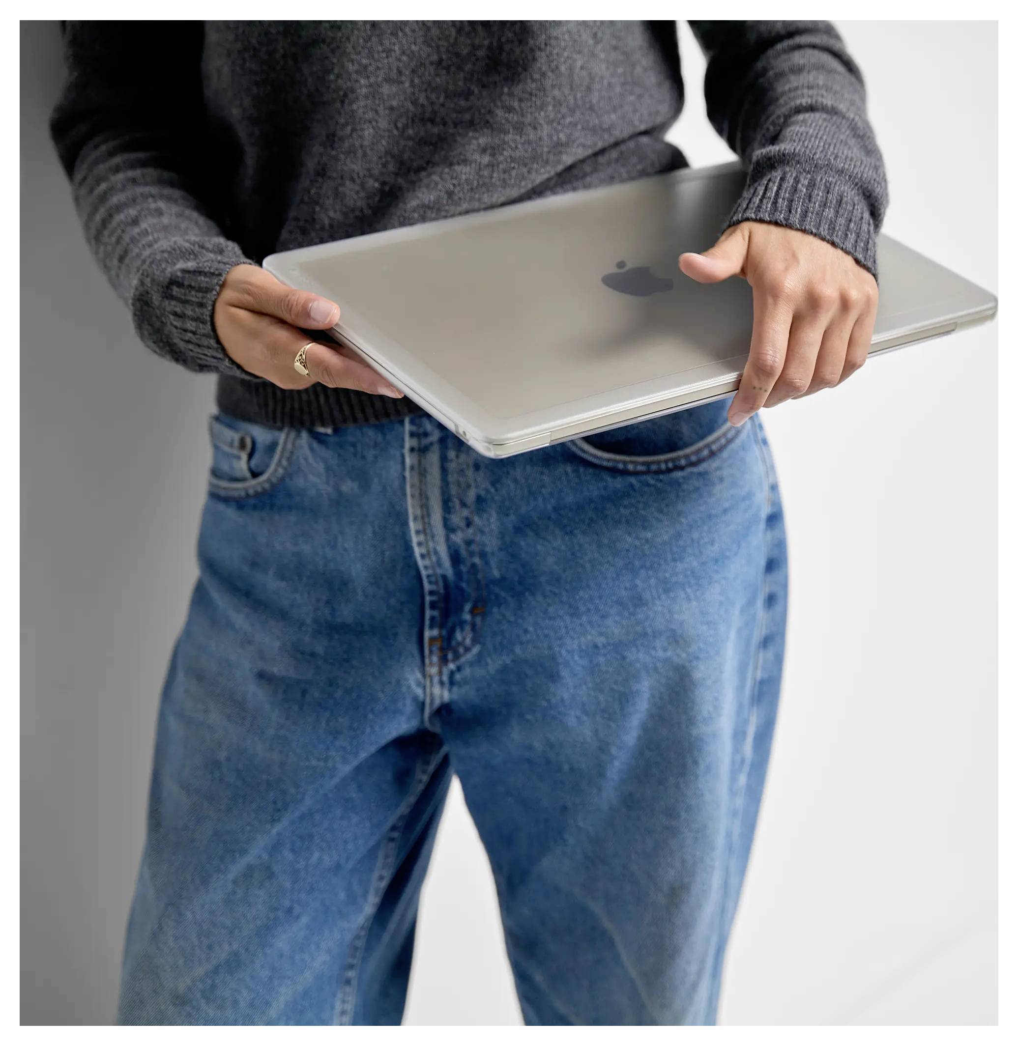 Een persoon in een grijze trui en blauwe spijkerbroek houdt een gesloten laptop voor een witte muur.