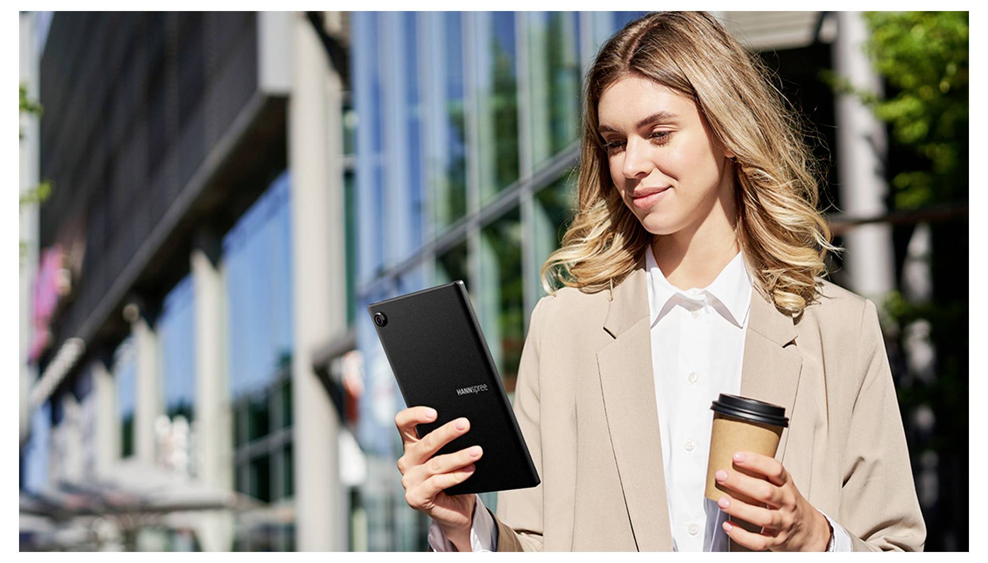 Een vrouw staat buiten een modern gebouw, een tablet en een koffiekopje vasthoudend, glimlachend en naar het scherm kijkend.