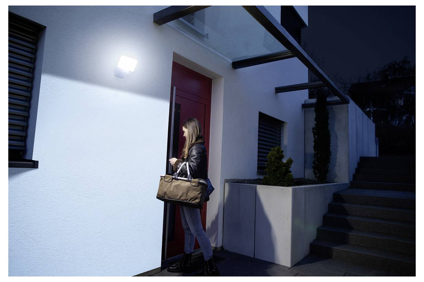 Een persoon betreedt 's avonds een modern, goed verlicht huis terwijl hij een bruine tas vasthoudt. De buitenkant heeft een strak design met trappen aan de rechterkant.