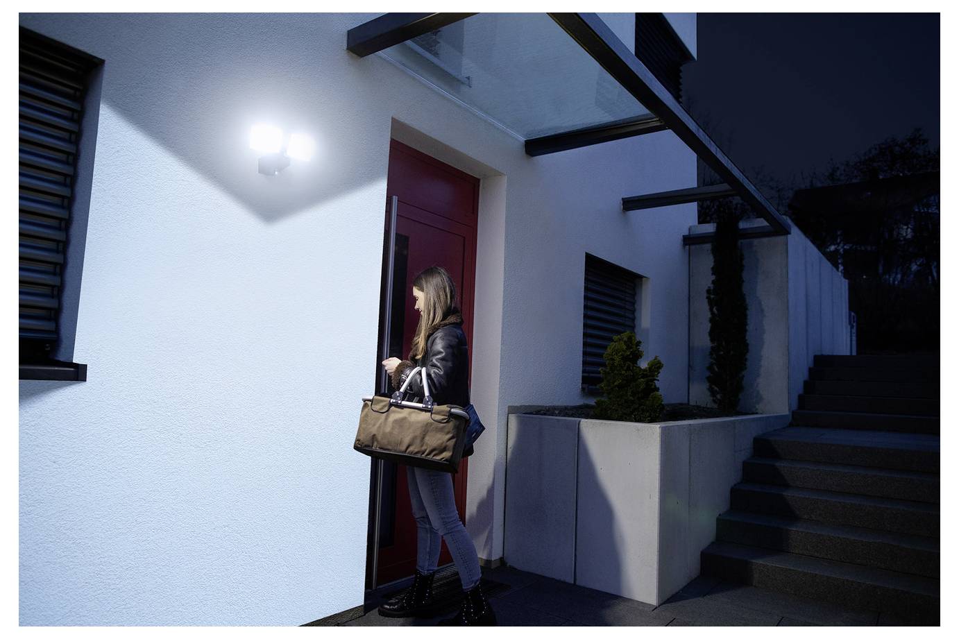 Een vrouw met een tas betreedt 's avonds een modern huis door een rode deur, verlicht door een helder, bewegingsgeactiveerd beveiligingslicht.