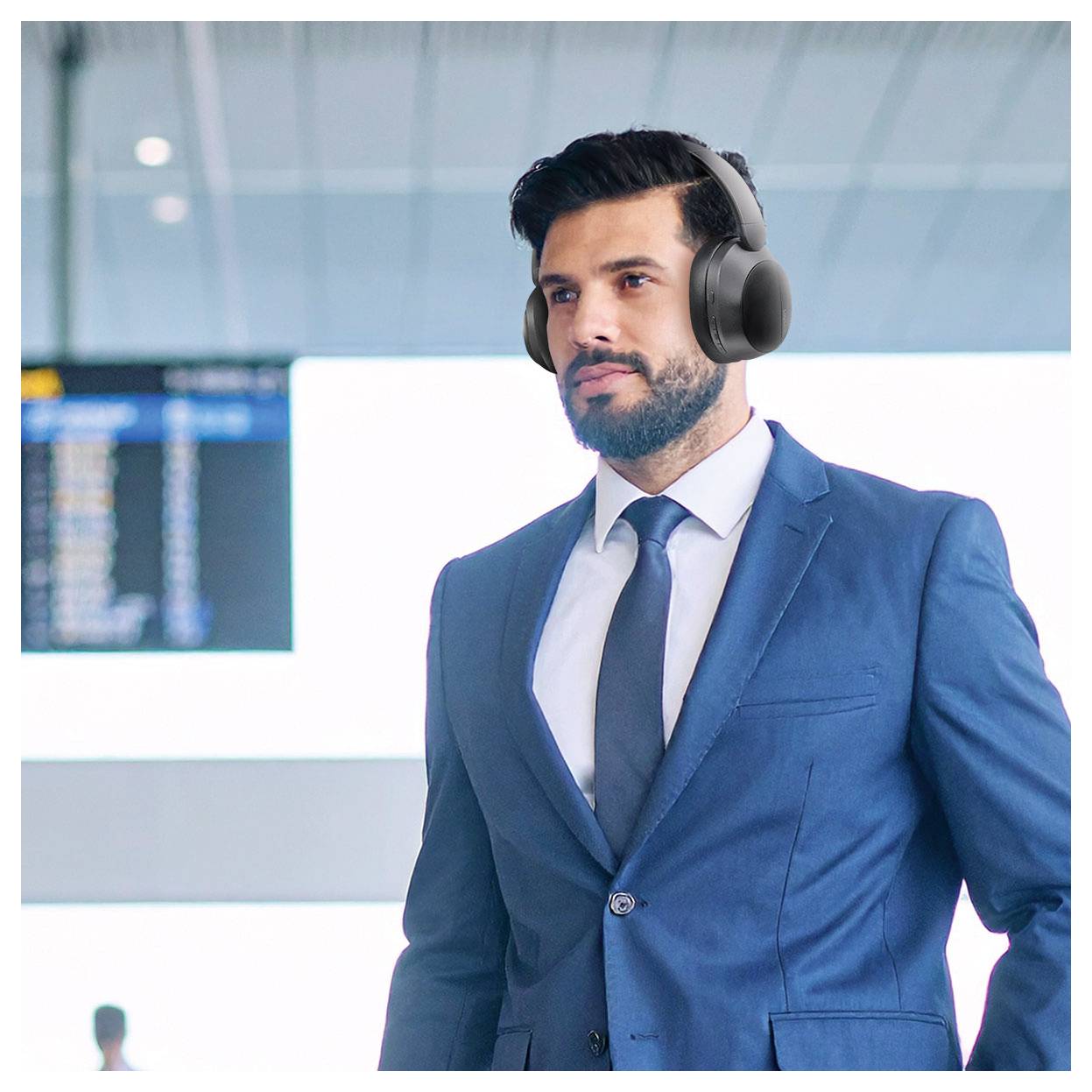 Een man in een blauw pak met hoofdtelefoons staat in een luchthaventerminaal, met een vluchtinformatiebord zichtbaar op de achtergrond.