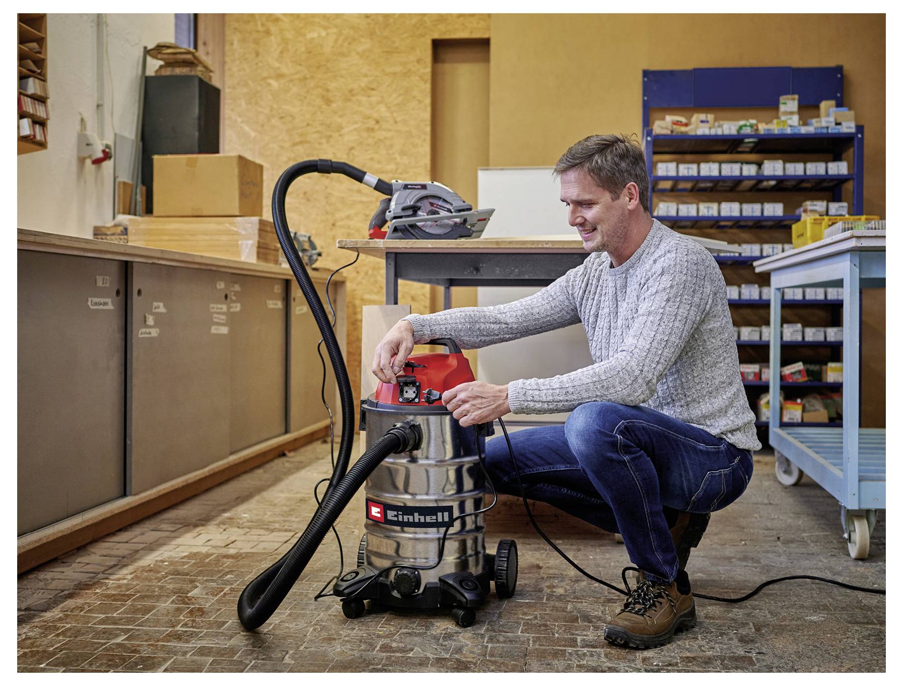 Man die een stofzuiger bedient in een werkplaats, omringd door gereedschappen en opslagplanken. Hij is de instellingen van de machine aan het aanpassen.