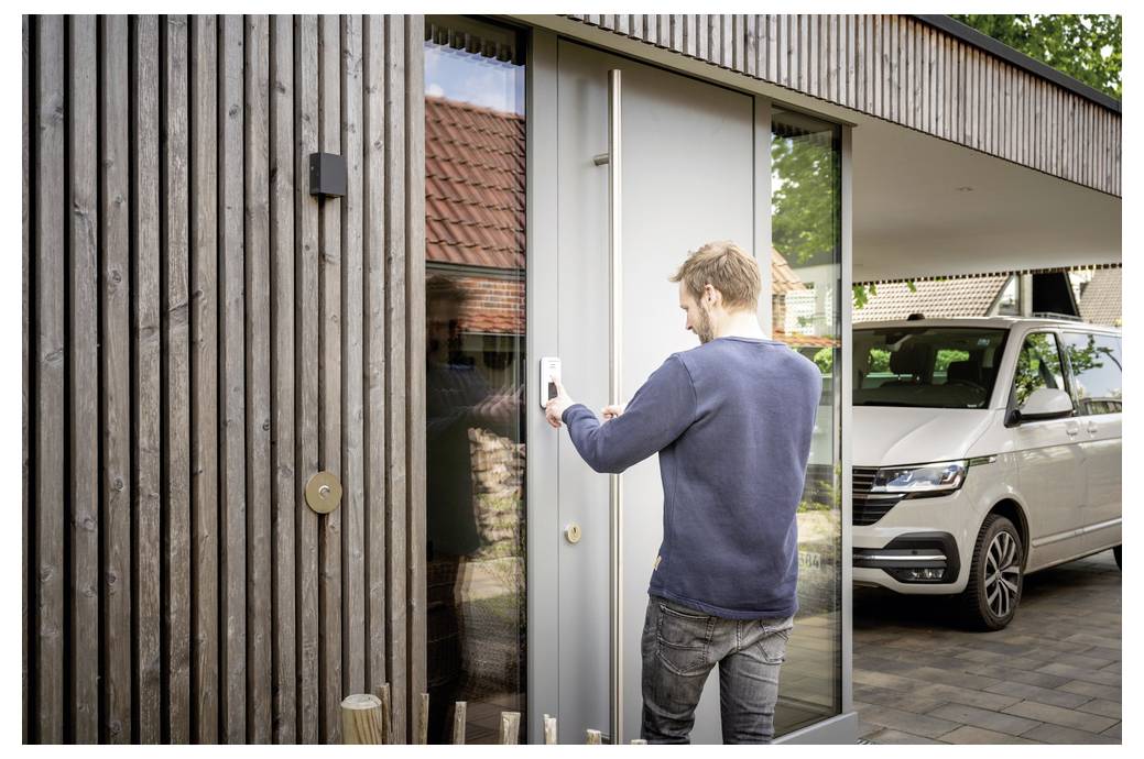 Een persoon die een smart lock gebruikt om een moderne woning met houten exterieur binnen te gaan. Een witte bestelwagen staat geparkeerd op de oprit.