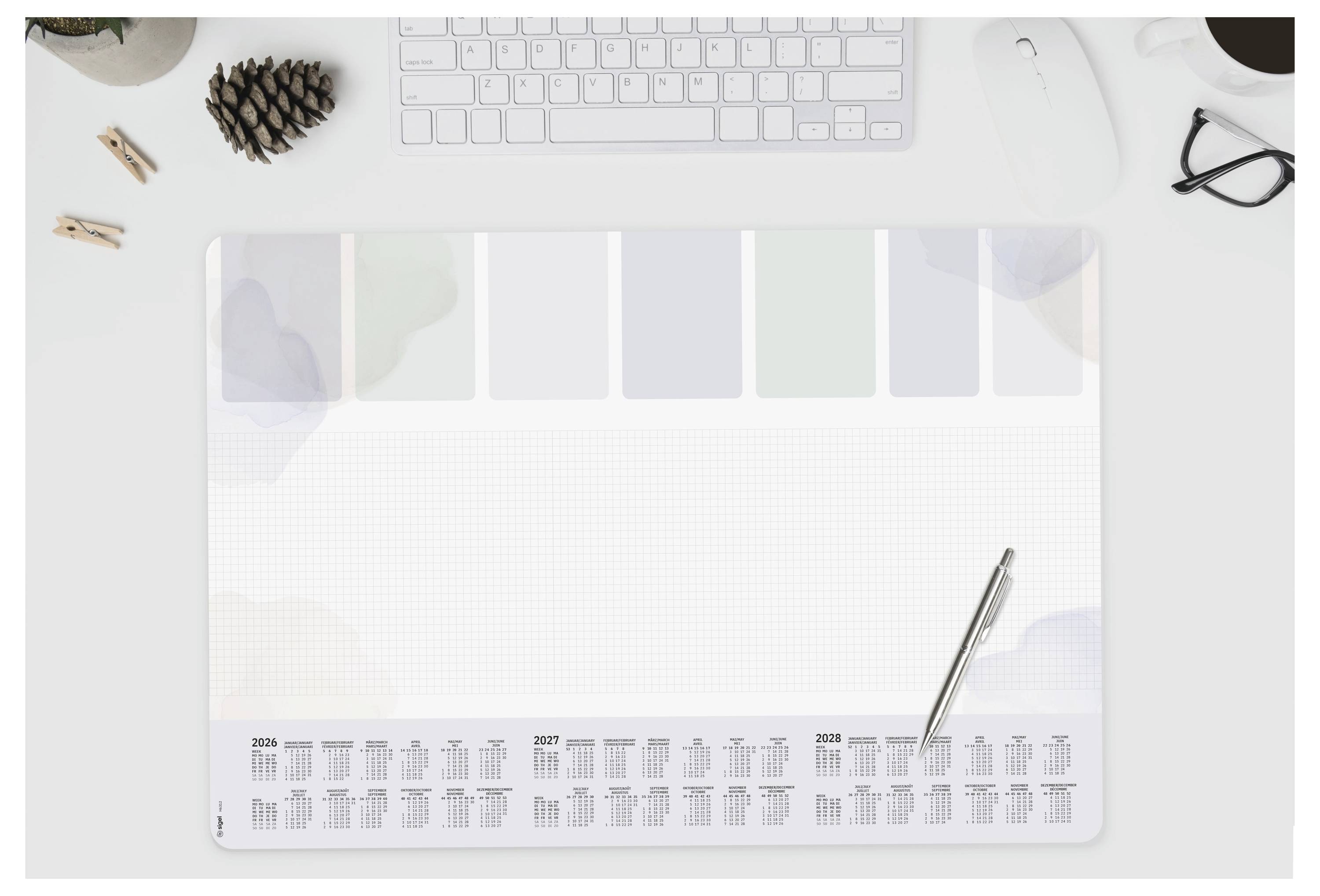 Een bureaukalender met een pen erop, een computertoetsenbord, muis, dennenappel, bril en een paar paperclips op een wit bureau.