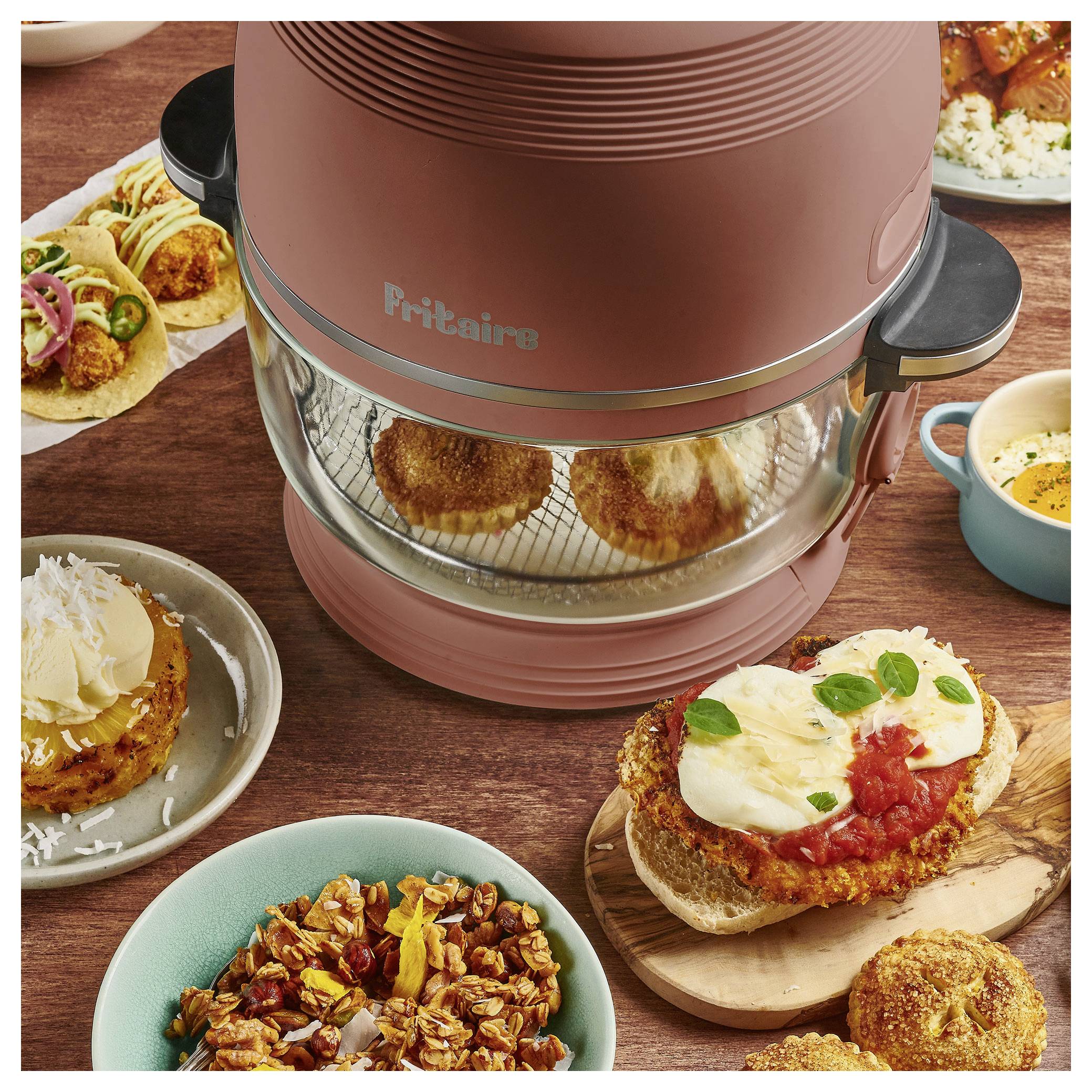 Een aanrecht in de keuken met een airfryer gevuld met voedsel. Eromheen borden met taco's, een hamburger bedekt met tomatensaus en kaas, en een kom met zaden.