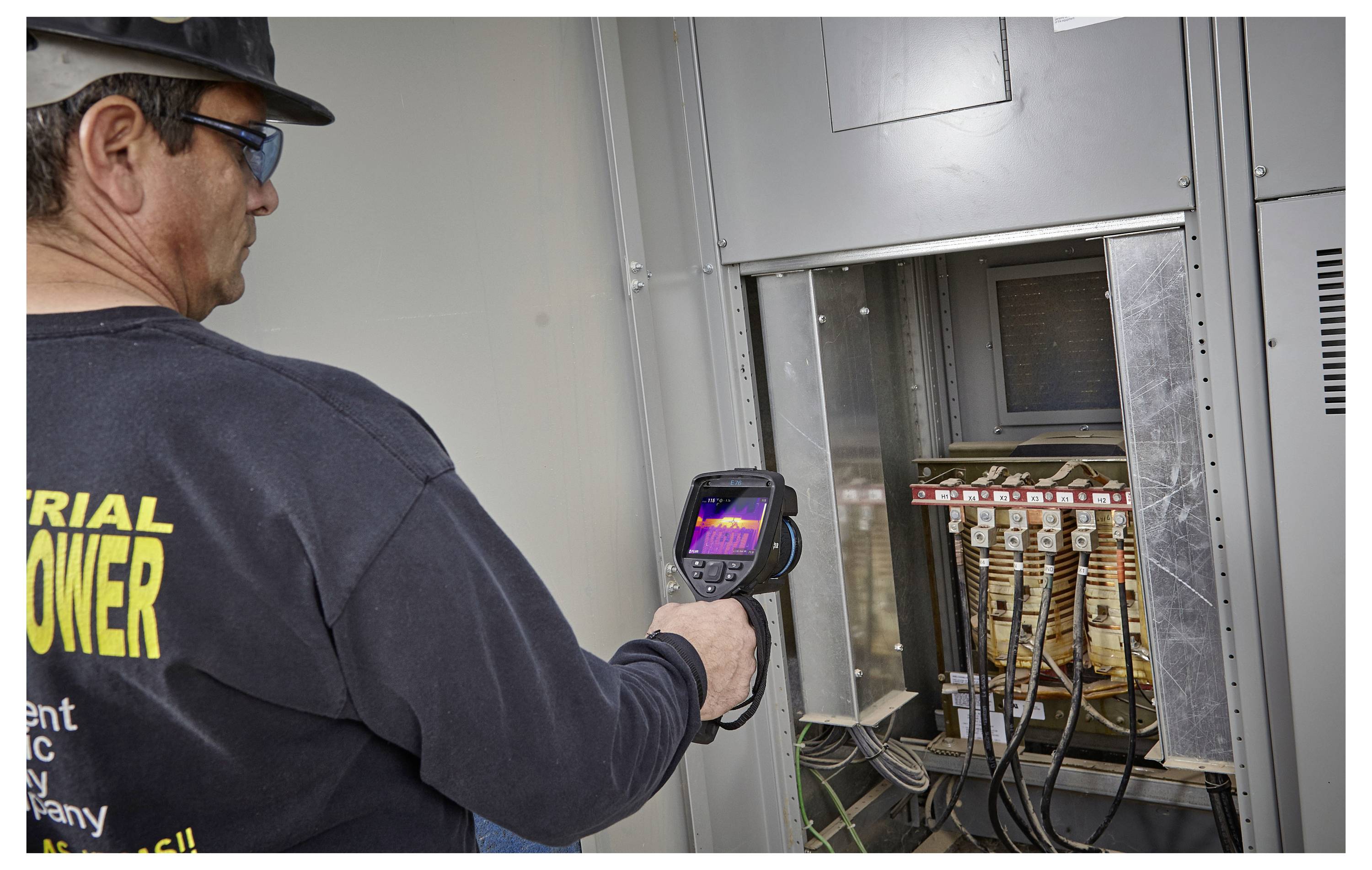 Een medewerker gebruikt een warmtebeeldcamera om elektrische bedrading in een paneel te inspecteren, gekleed in veiligheidsuitrusting in een industriële omgeving.