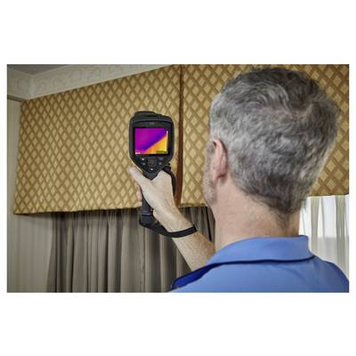 Een persoon die een warmtebeeldcamera gebruikt om een kamer met gedessineerde gordijnen te inspecteren, waarschijnlijk om warmteverlies of isolatieproblemen te controleren.