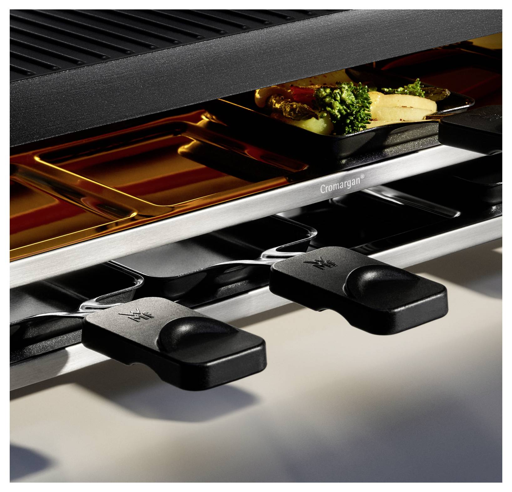 Close-up van een meerlaagse grill met een bakplaat waarop groenten en vis worden bereid. Het zwarte, geprofileerde oppervlak en het met 'Cromargan' gelabelde metalen bakblik zijn zichtbaar.