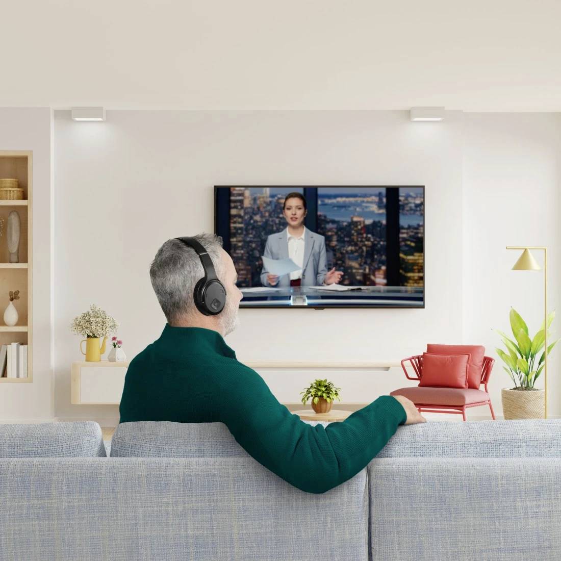 Een persoon in een groene trui kijkt naar een nieuwsuitzending op televisie terwijl hij hoofdtelefoons draagt, gezeten in een moderne woonkamer.