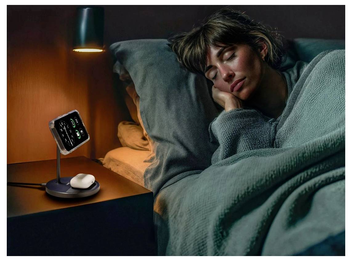 Een vrouw slaapt in bed onder een groene deken. Een mobiele telefoon op een nachtkastje toont gezondheidsmonitoringsgegevens onder een kleine lamp.