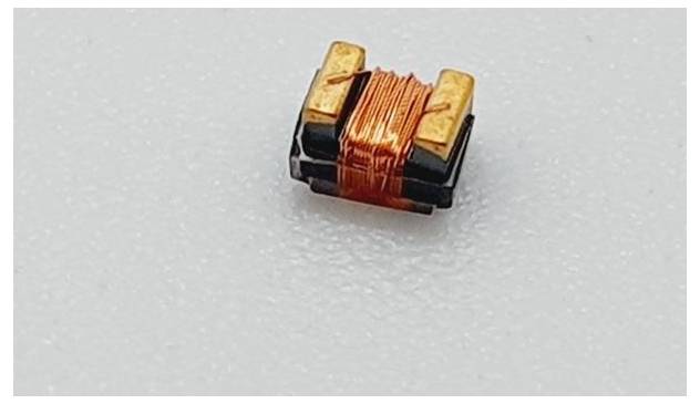 Een klein elektronisch onderdeel met koperdraadspoelen wordt geplaatst op een wit oppervlak, waarschijnlijk een inductor of transformator die wordt gebruikt in elektronische schakelingen.