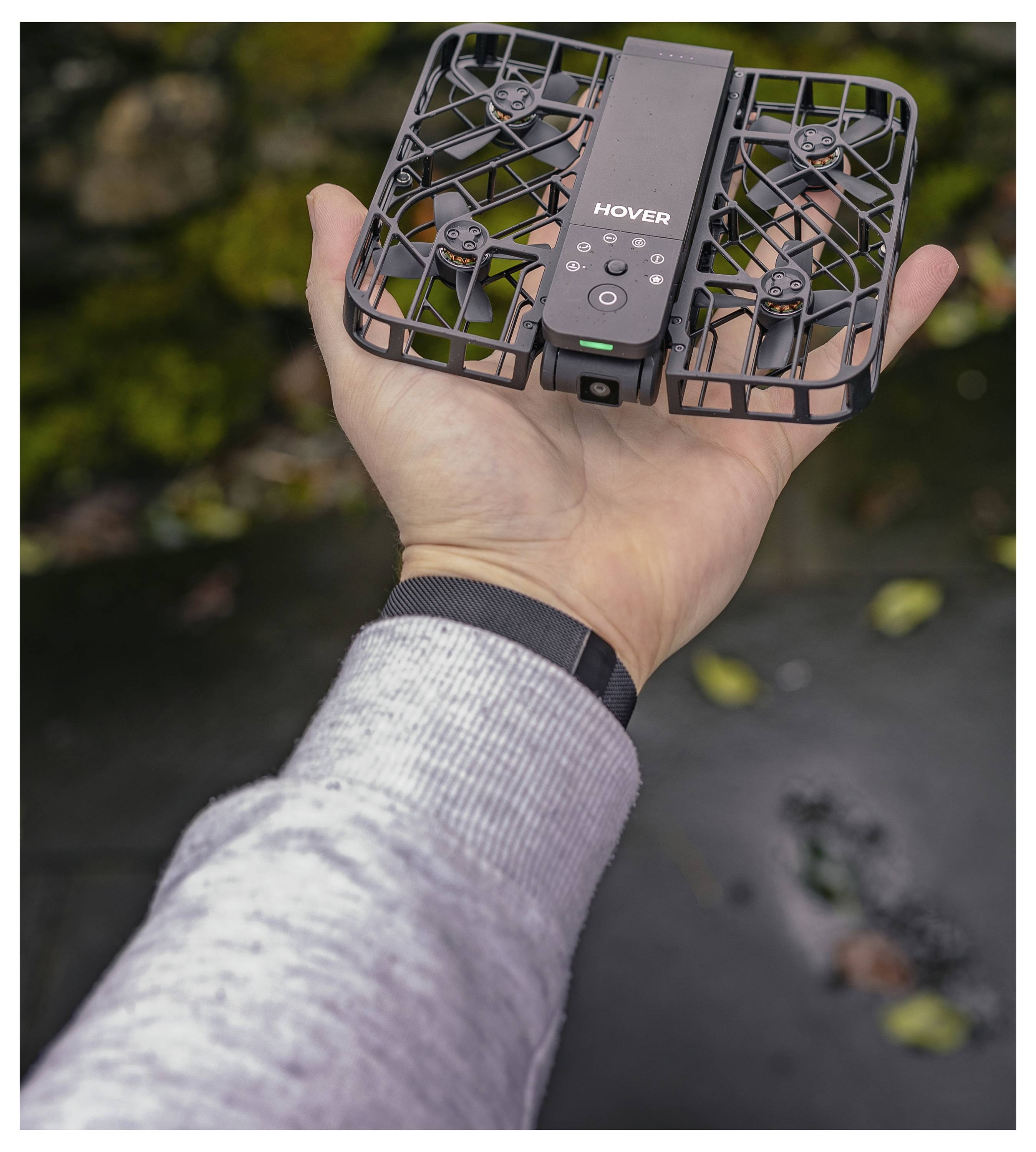 Een hand van een persoon die een kleine, zwarte, vierkante drone vasthoudt met het label 'HOVER', uitgerust met beschermende roosters rond de propellers.