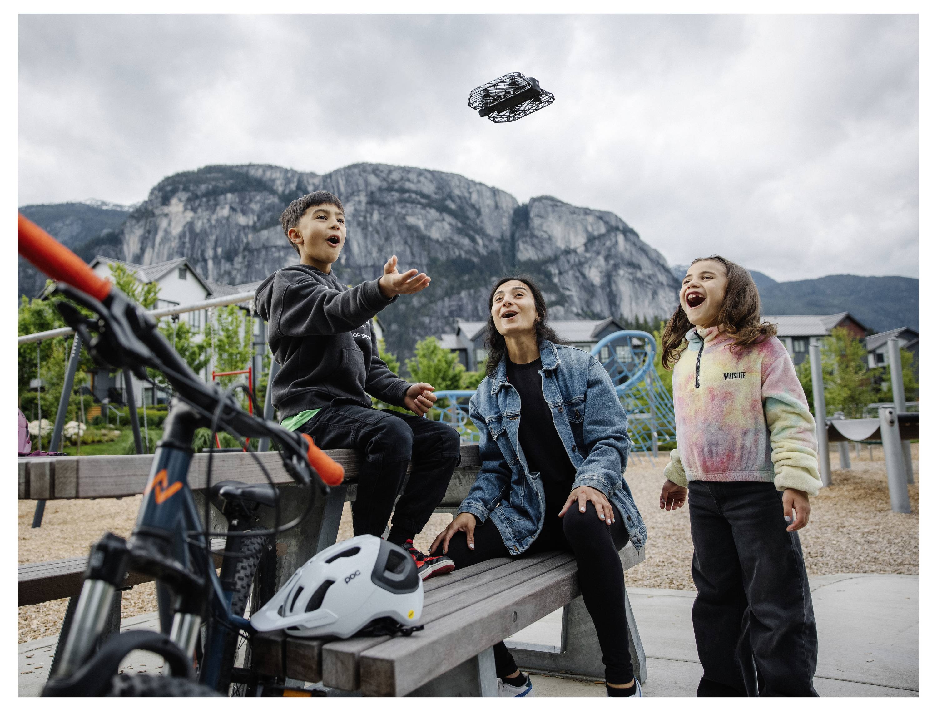 Een vrouw en twee kinderen kijken vrolijk naar een kleine drone die in een park vliegt. Een fiets en helm liggen in de buurt, met bergen op de achtergrond.