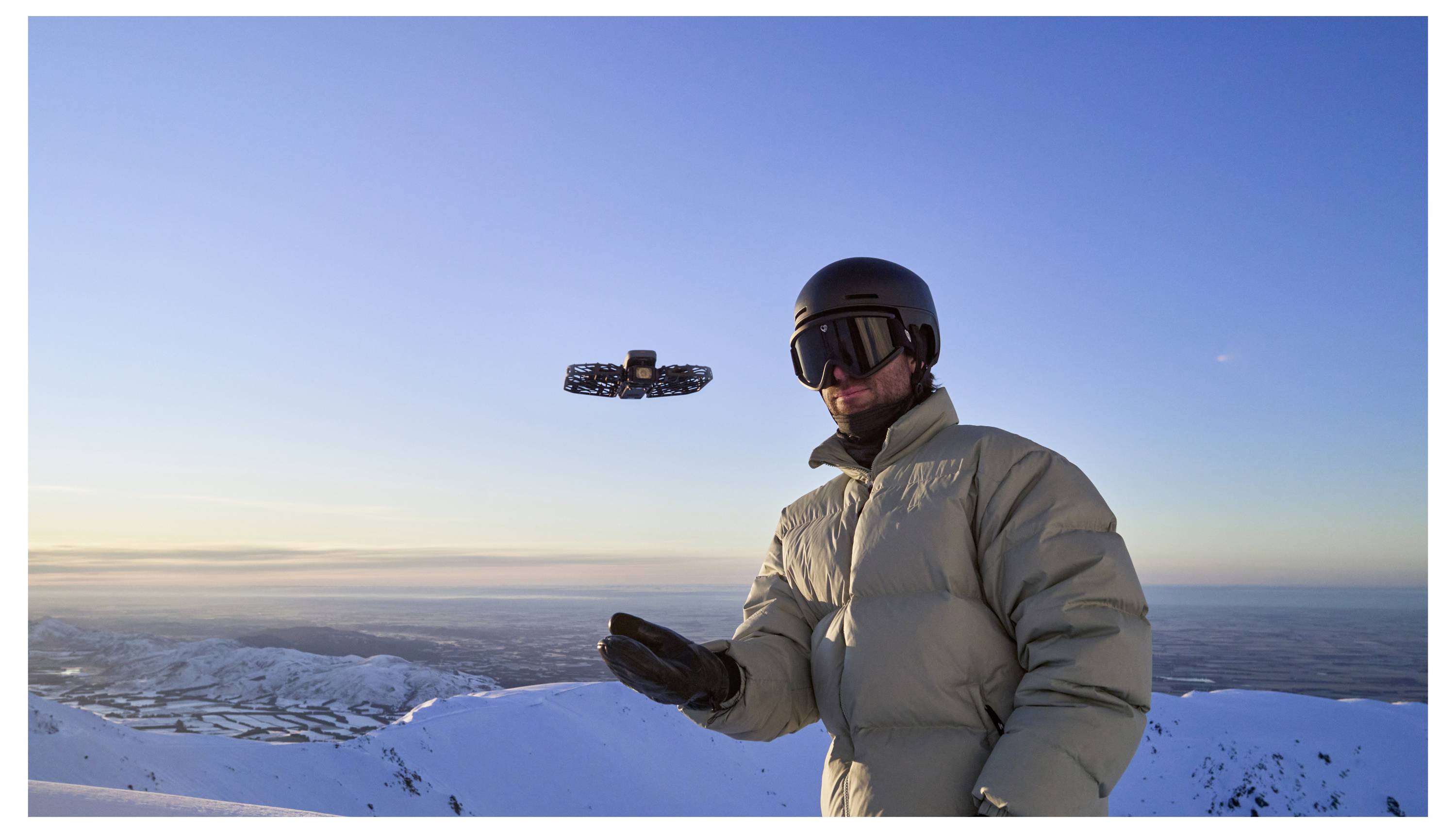 Een persoon in winteruitrusting die een kleine drone bestuurt in een besneeuwde berglandschap tijdens zonsondergang. De drone zweeft voor hem.