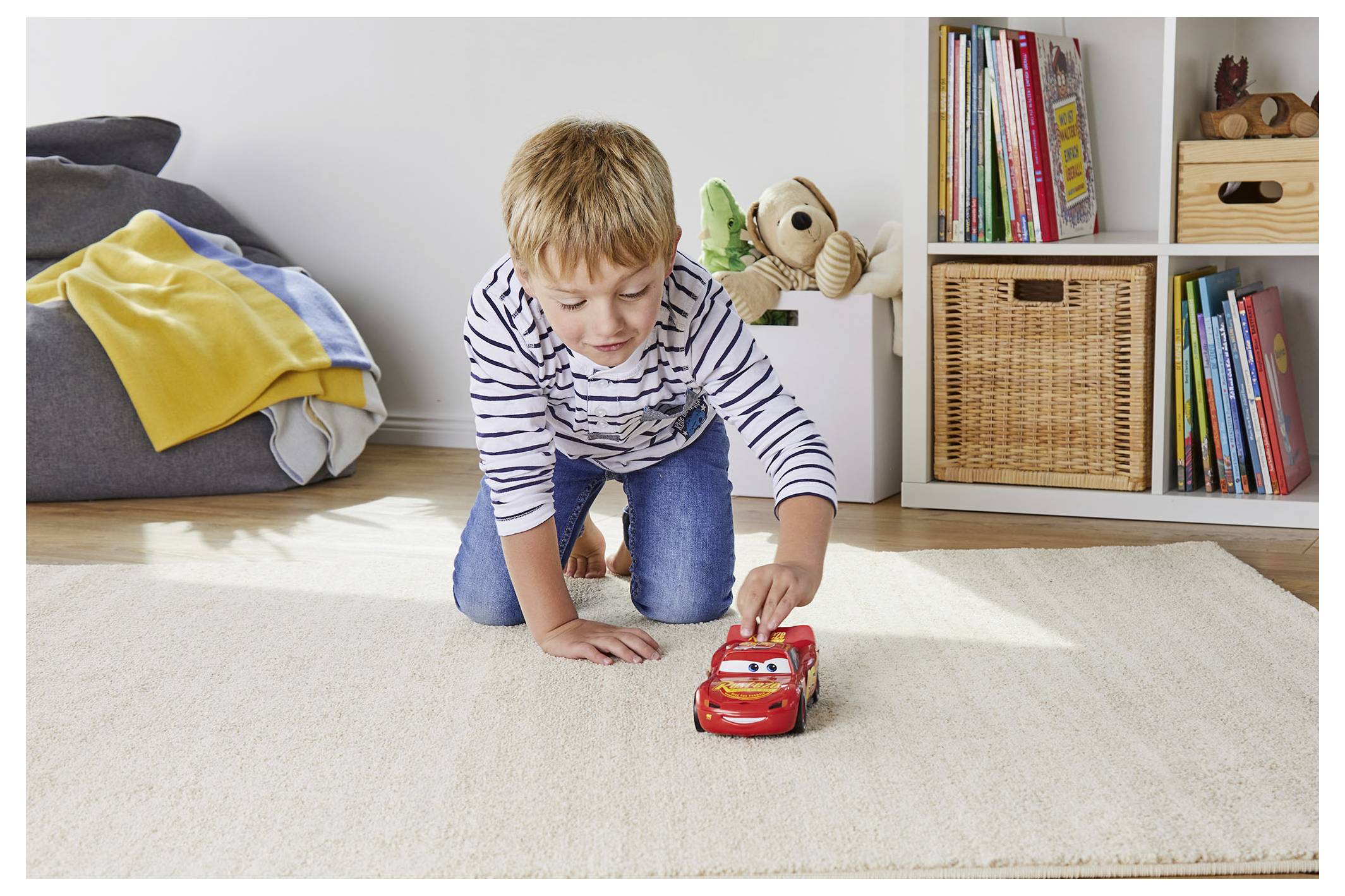 Een kind speelt op een vloerkleed met een speelgoedauto in een gezellige kamer met planken vol boeken, een gevlochten mand en een stapel dekens in de buurt.