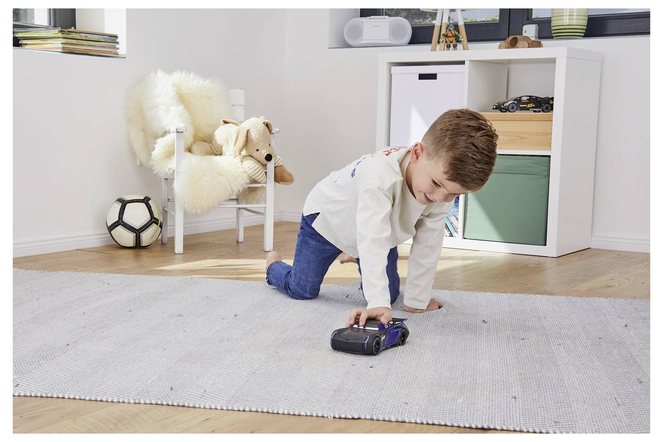 Een kind speelt met een speelgoedauto op een vloerkleed in een lichte kamer. Dichtbij staat een stoel met een knuffeldier en een voetbal, en rekken zijn op de achtergrond.