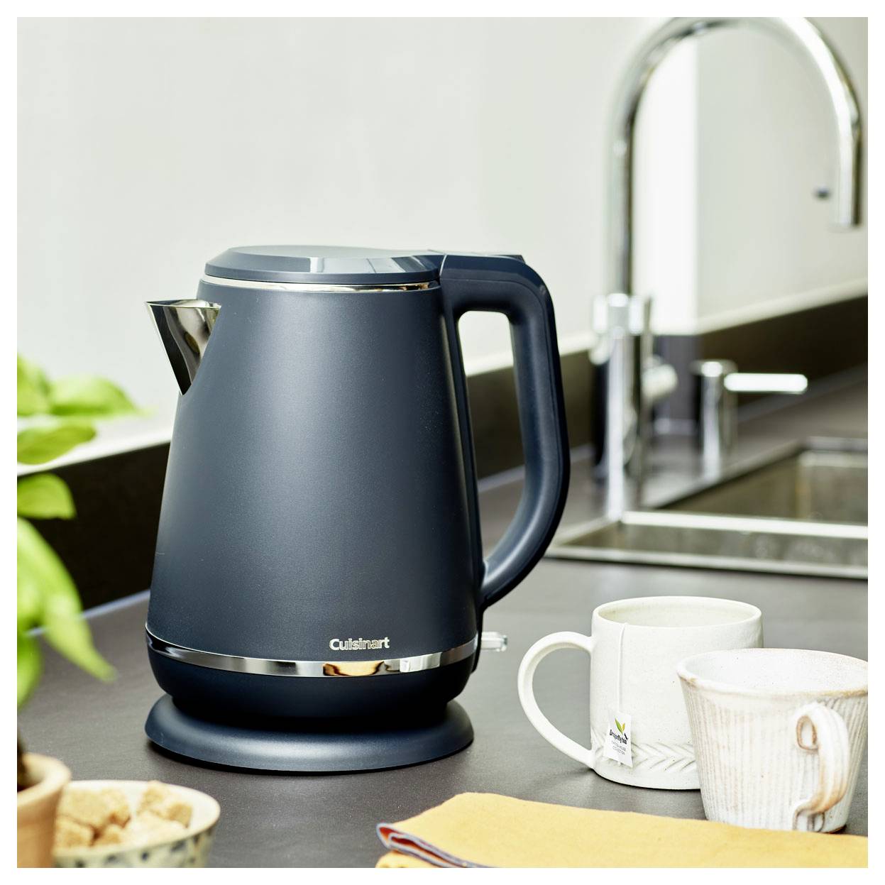 Een moderne zwarte elektrische waterkoker op een aanrecht naast twee keramische mokken en een groene plant bij een gootsteen.