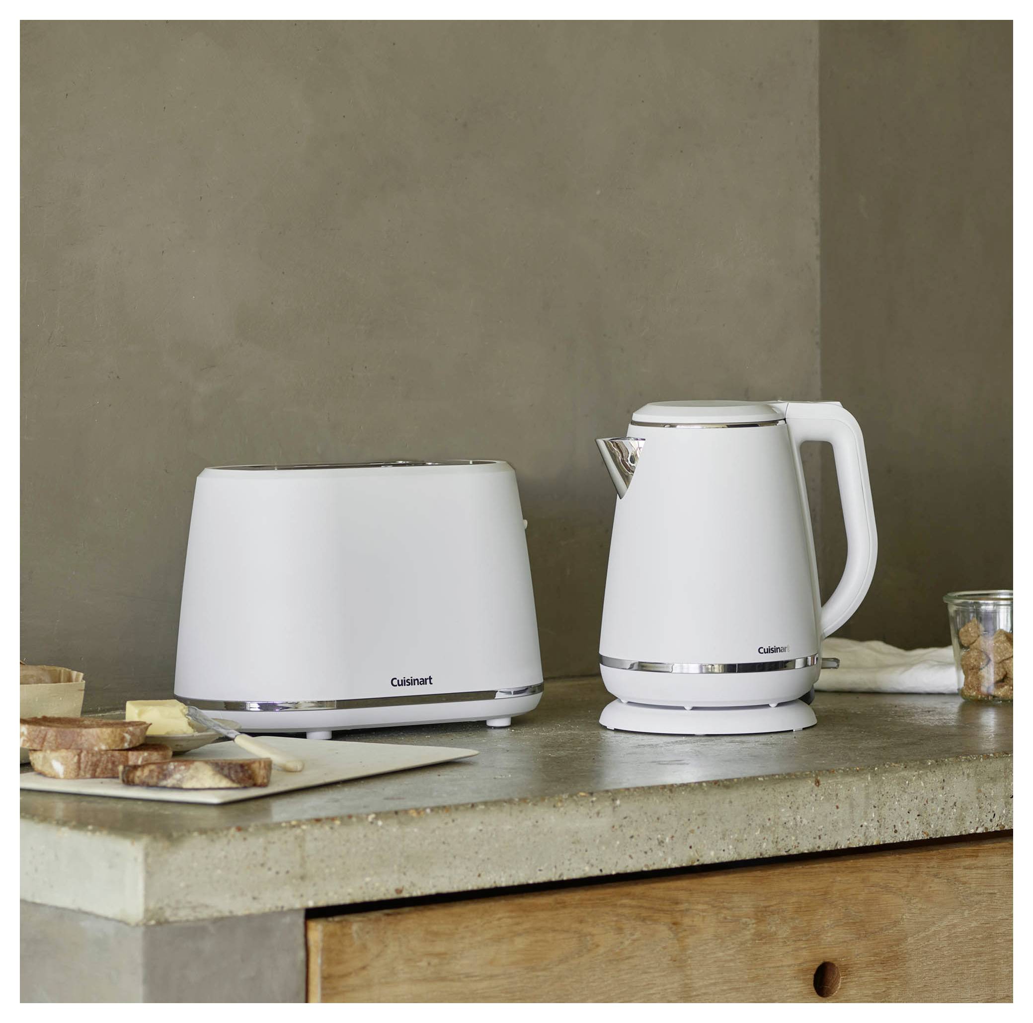 Een witte broodrooster en waterkoker staan op een houten aanrechtblad, met sneetjes brood ernaast.