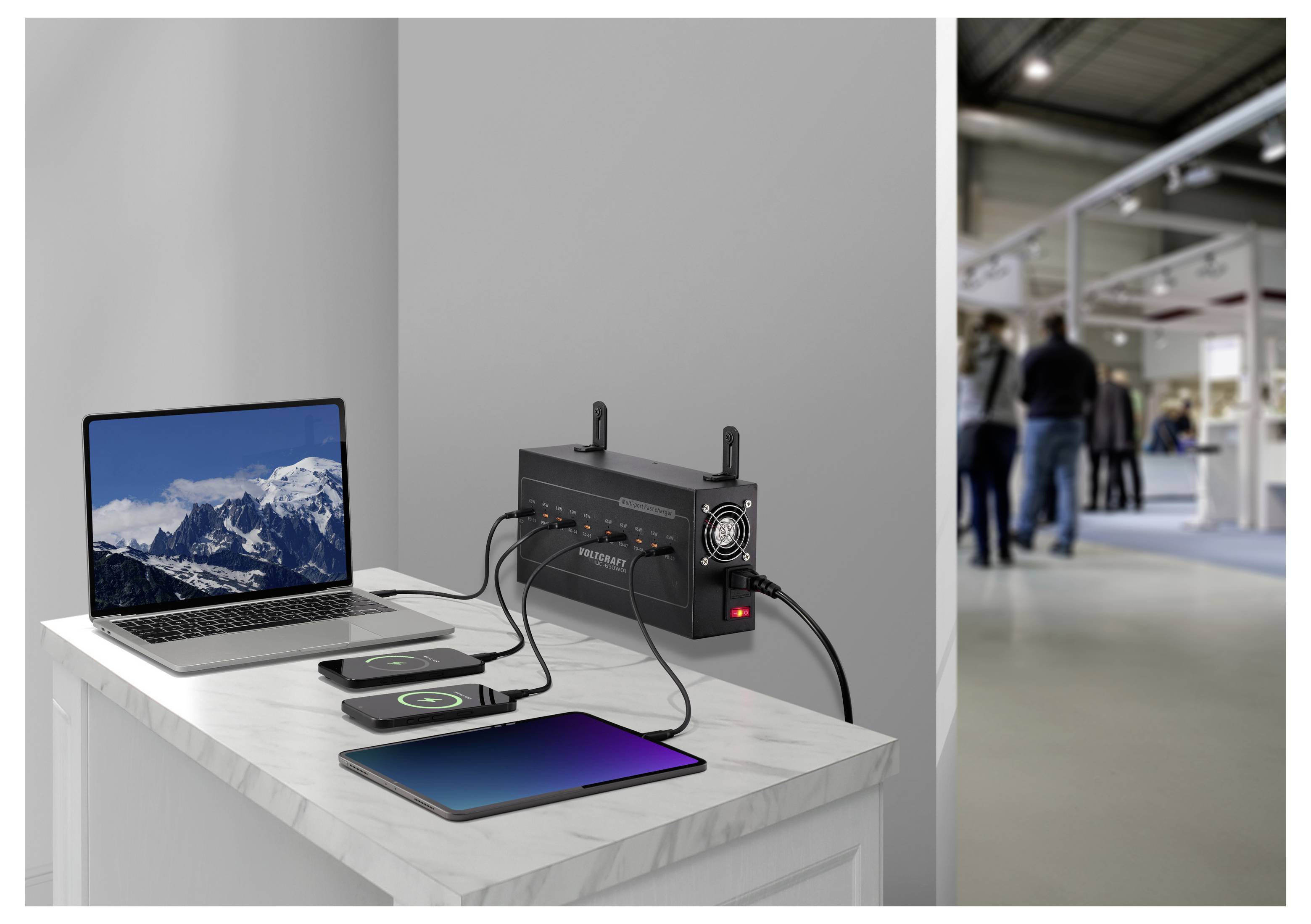Draagbaar stromaggregraat dat een laptop, smartphone en tablet oplaadt op een witte tafel, met een vervaagde vloer van een beurs op de achtergrond.