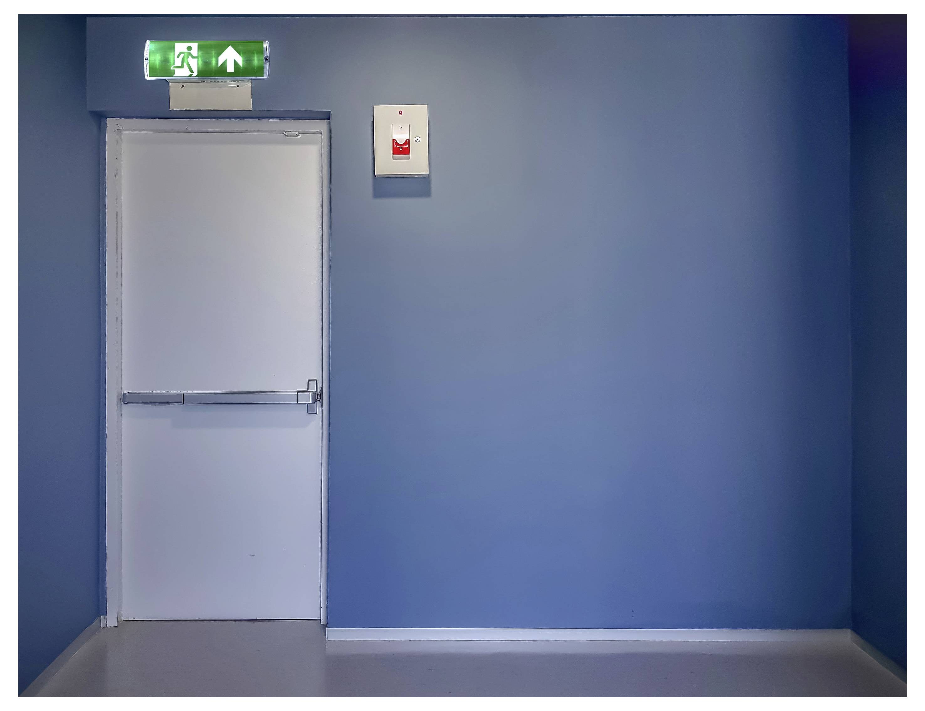 'Afbeelding van een gesloten witte nooduitgangdeur in een kamer met blauwe muren, met een verlicht groen uitgangsbordje erboven, waarop een persoon die rent is afgebeeld.'