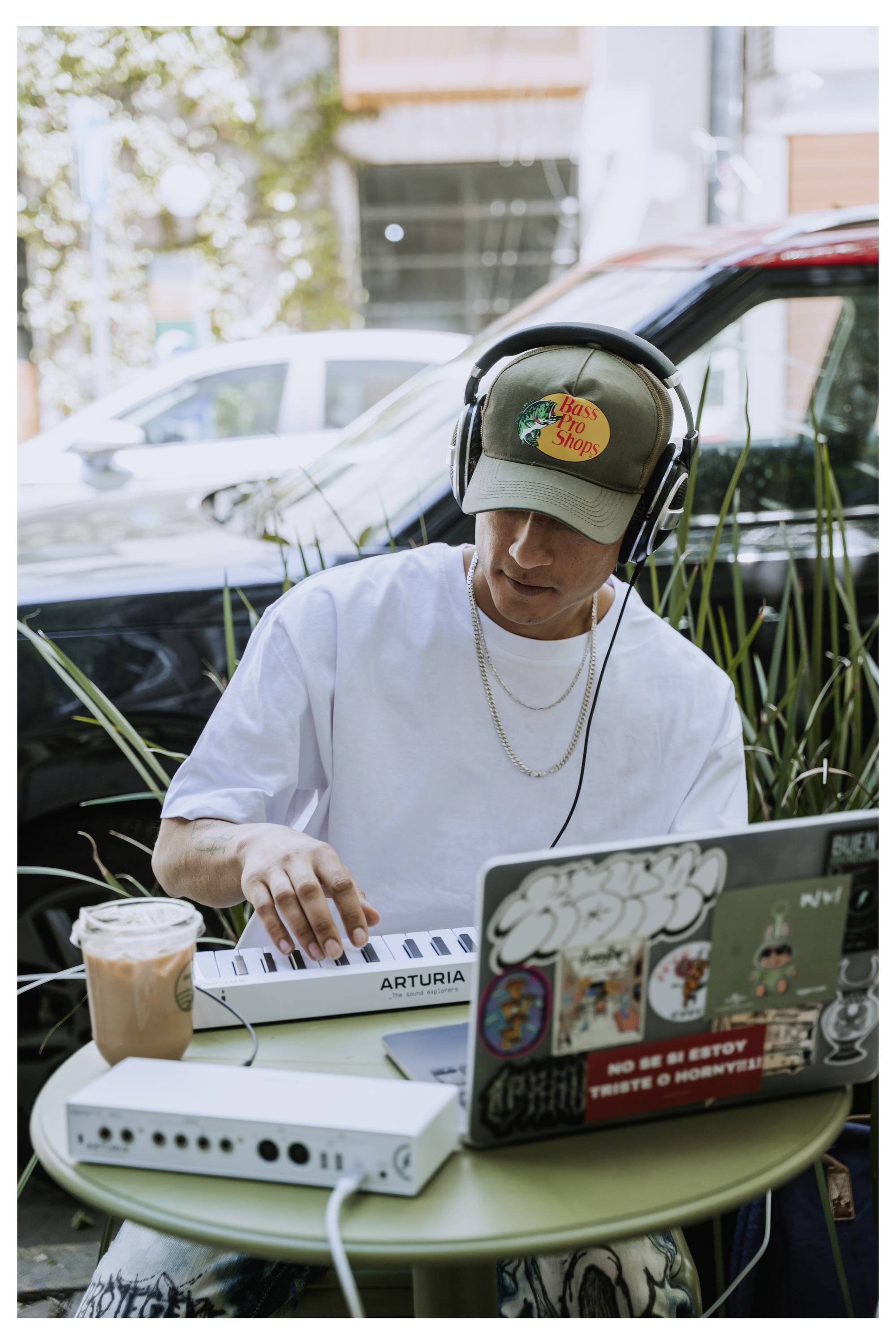 Een persoon met een koptelefoon werkt met muziekapparatuur en een laptop buiten, omringd door planten en auto's.