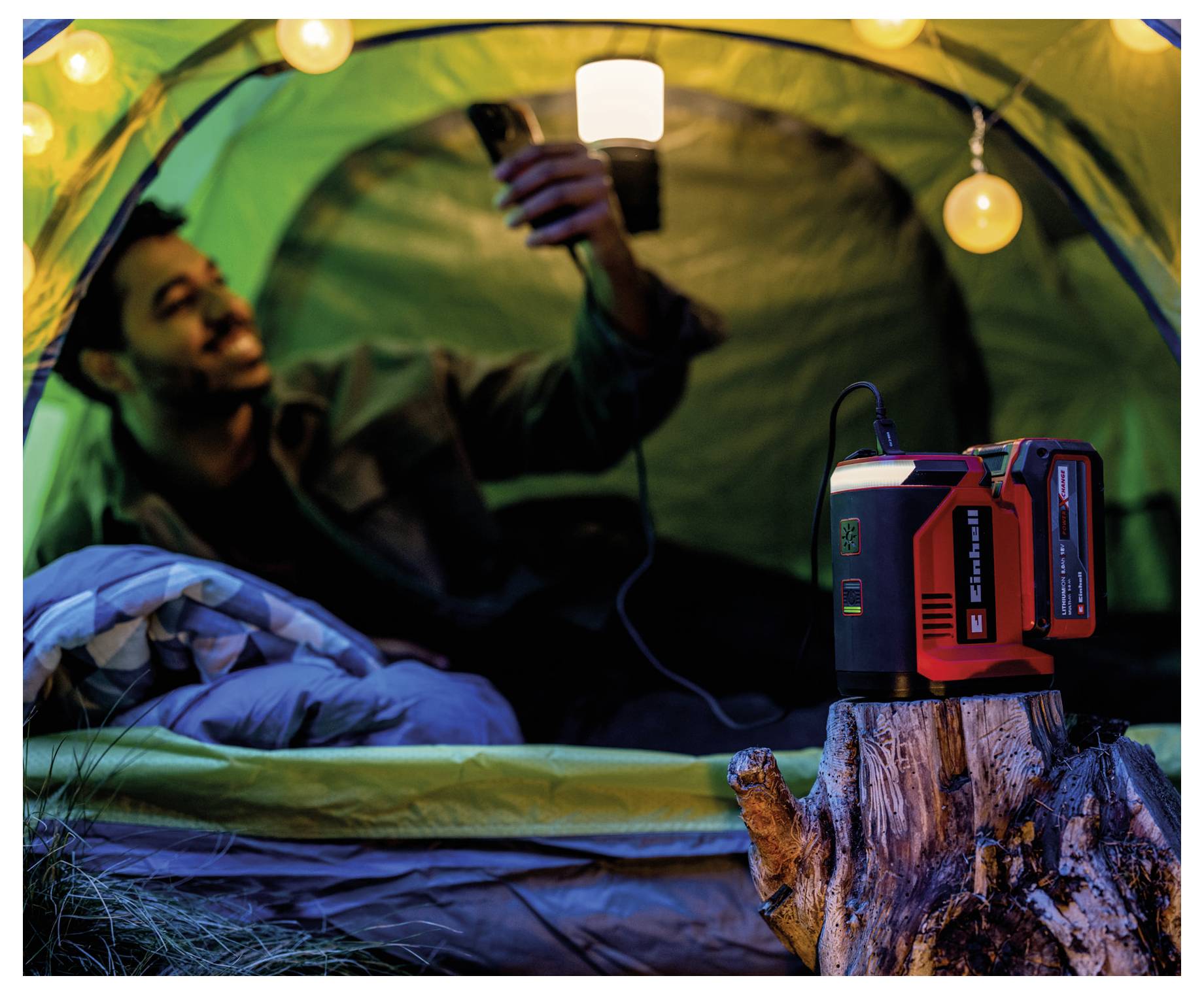 Een persoon ontspant zich in een verlichte tent terwijl hij een mobiele telefoon gebruikt, met een draagbaar stroomstation op een boomstronk buiten, omringd door lichten.
