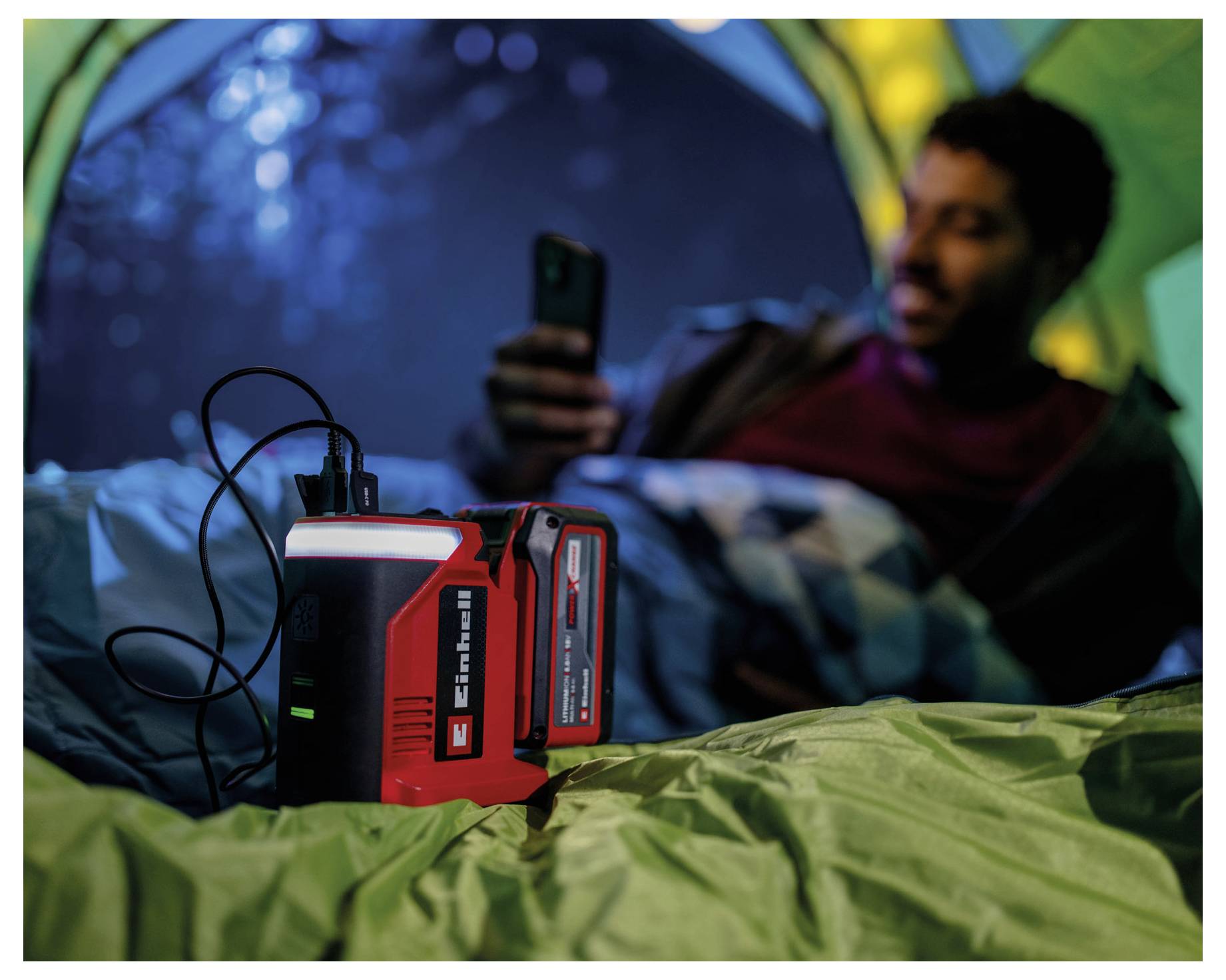 Een persoon in een tent gebruikt een smartphone die is aangesloten op een rode draagbare powerbank, wat wijst op het gebruik van kampeer- en technologie-uitrusting.