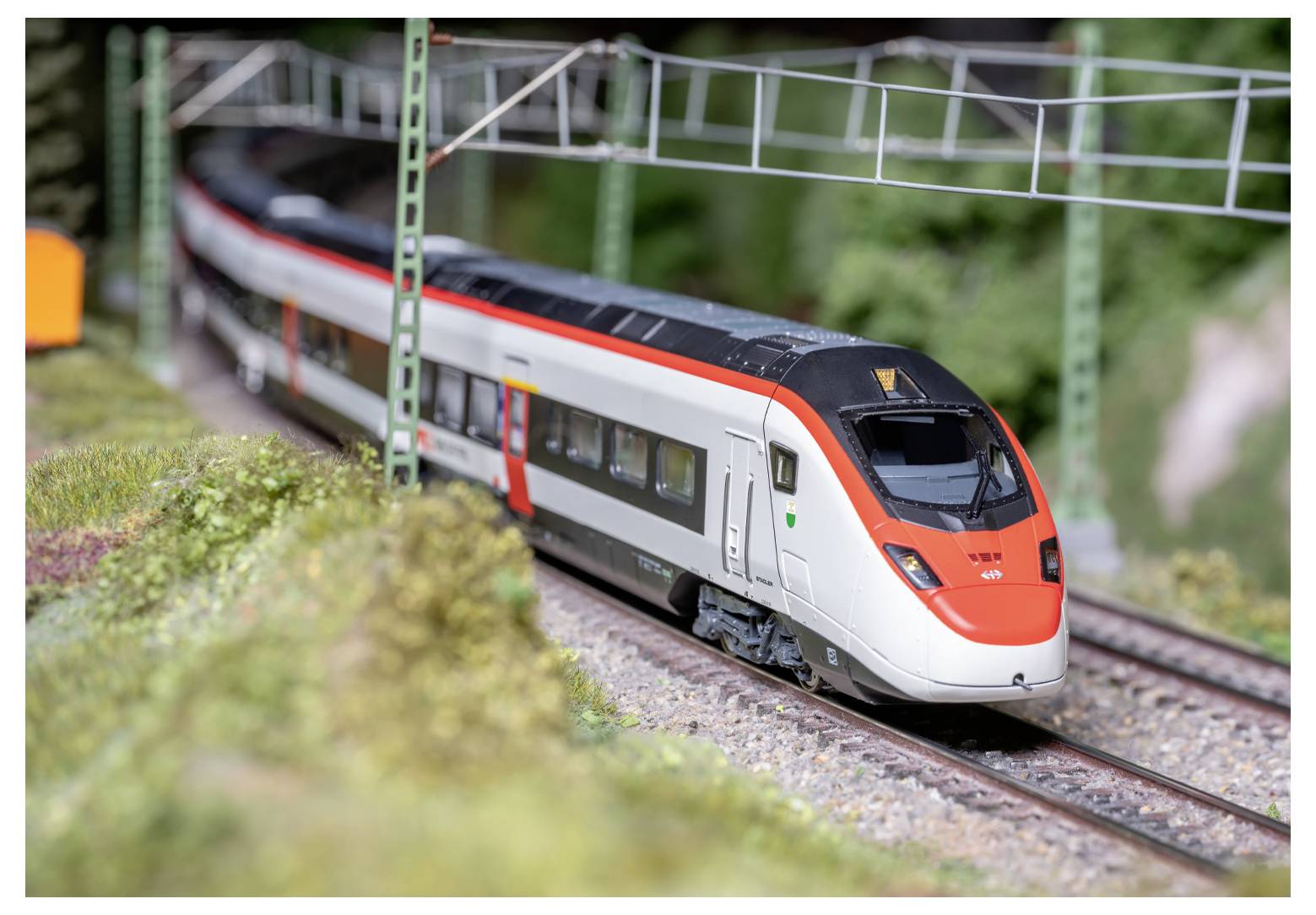 Een modeltreintje met rode en witte kleuren rijdt over rails onder groene metalen portaalbruggen, omgeven door gedetailleerd miniatuurlandschap.