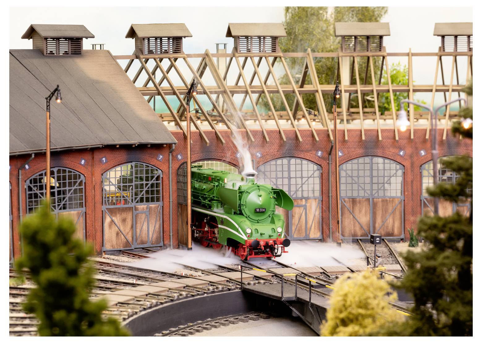 Een modelmatige groene stoomlocomotief komt tevoorschijn uit een locomotievenloods op een spoor, omgeven door bomen en gedetailleerde miniatuurlandschappen.