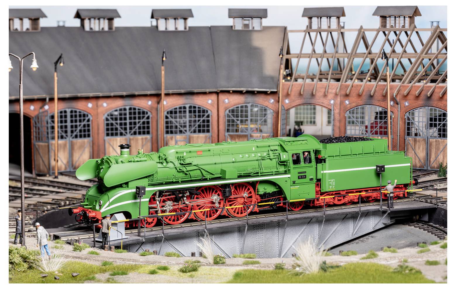 Een model van een groene stoomlocomotief met rode wielen op een draaischijf voor een locomotievenloods. Een persoon staat links.