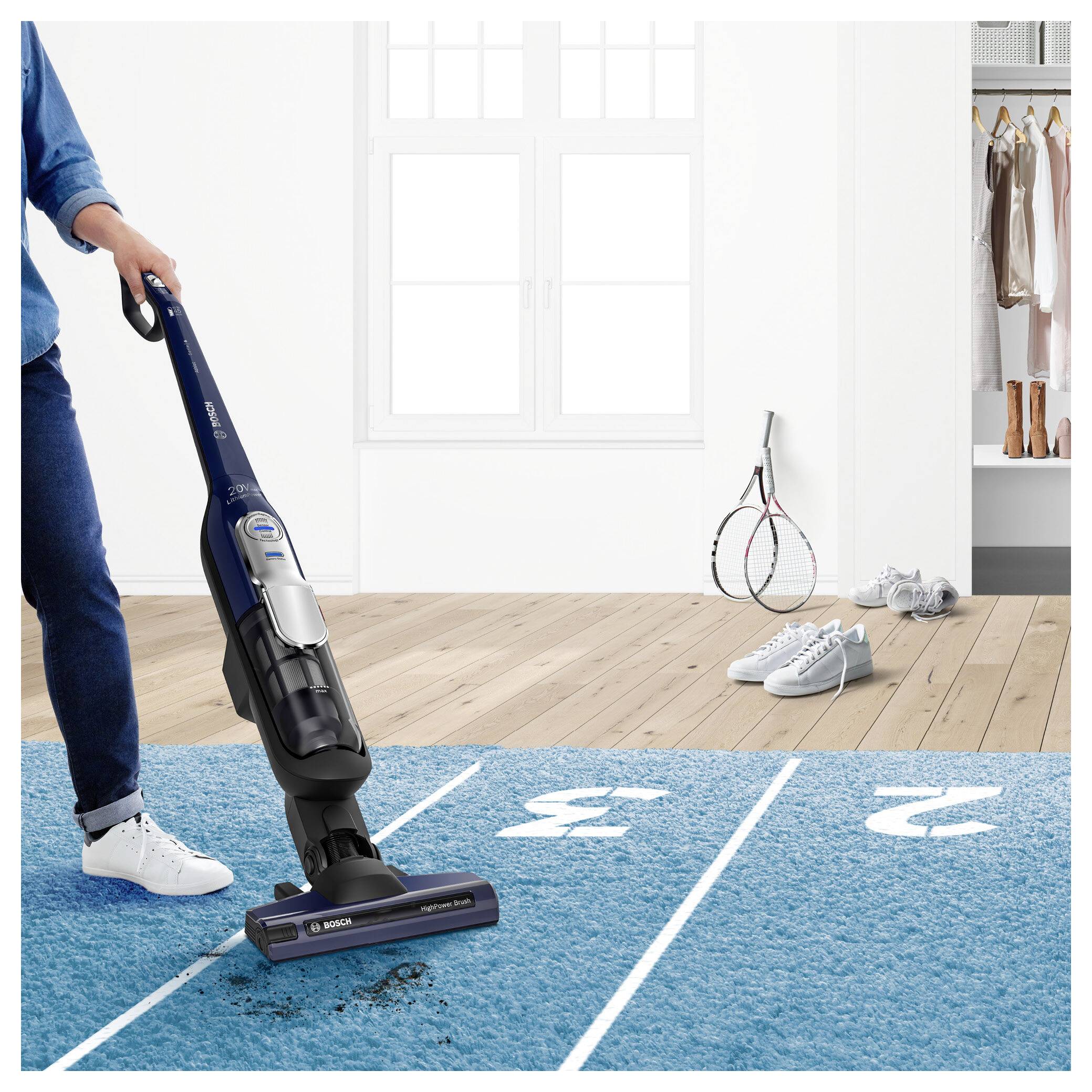 Een persoon stofzuigt een vloerkleed dat beschilderd is als een 100-meter racebaan, terwijl tennisuitrusting naast zich op de houten vloer staat.