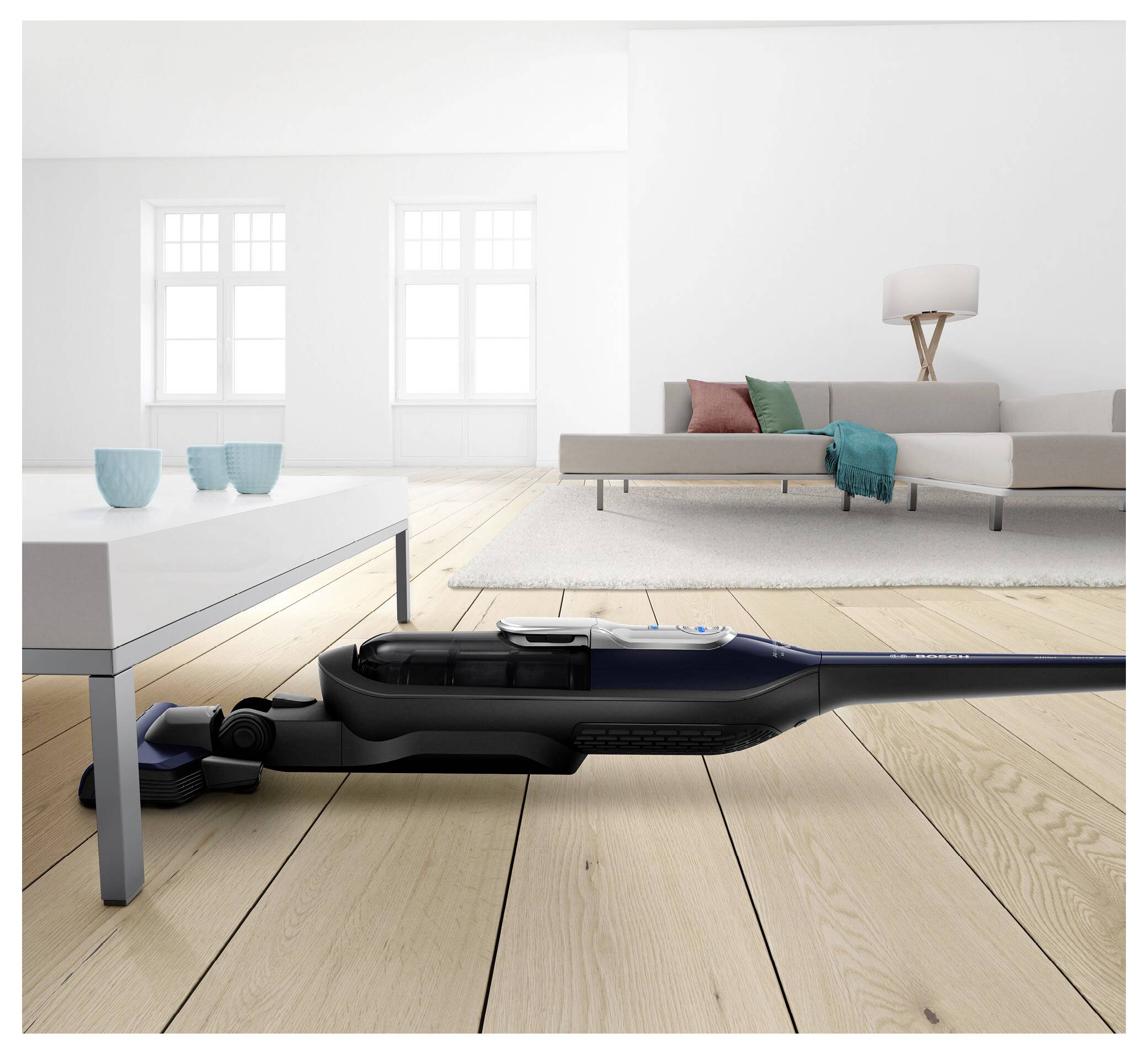 Een stofzuigerligt plat om onder een witte salontafel schoon te maken in een lichte woonkamer met een bank en lamp op de achtergrond.