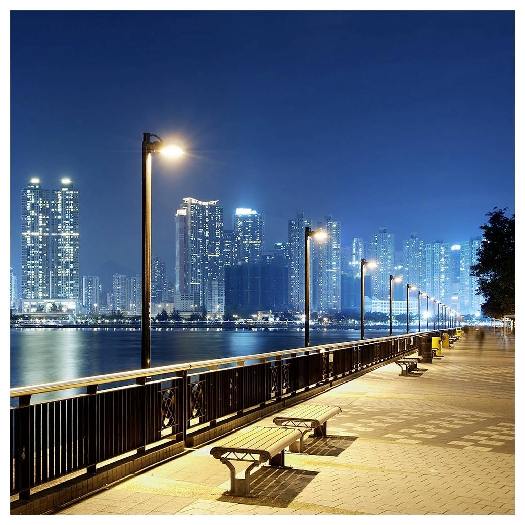 Een stadsgezicht 's nachts met een waterkantpromenade, straatverlichting, bankjes en een zicht op verlichte wolkenkrabbers aan de overkant van het water.