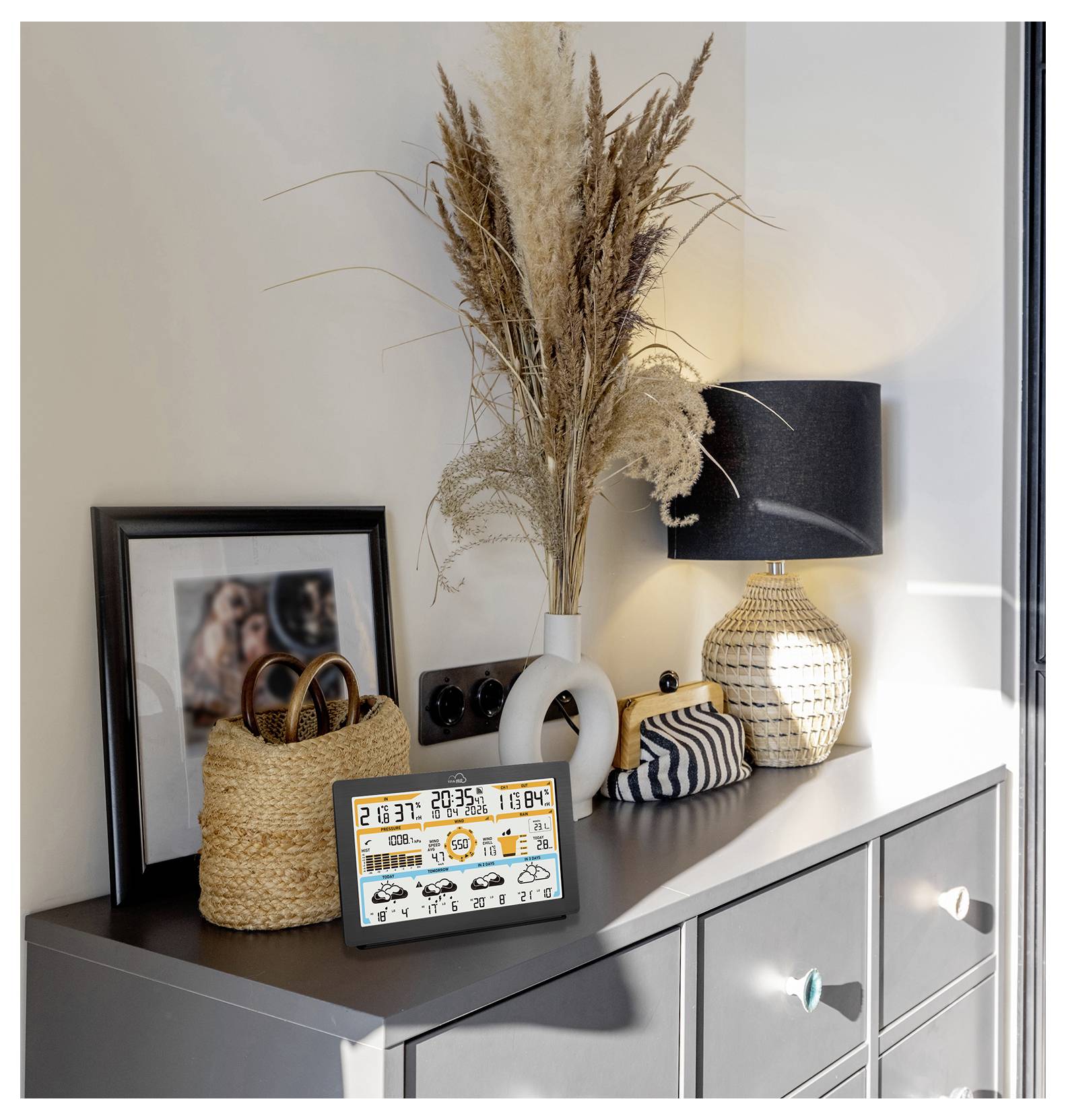 Een dressoir met een decoratieve lamp, een ingelijste foto, rieten manden en een digitaal display dat de tijd, datum, het weer en de temperatuur toont.