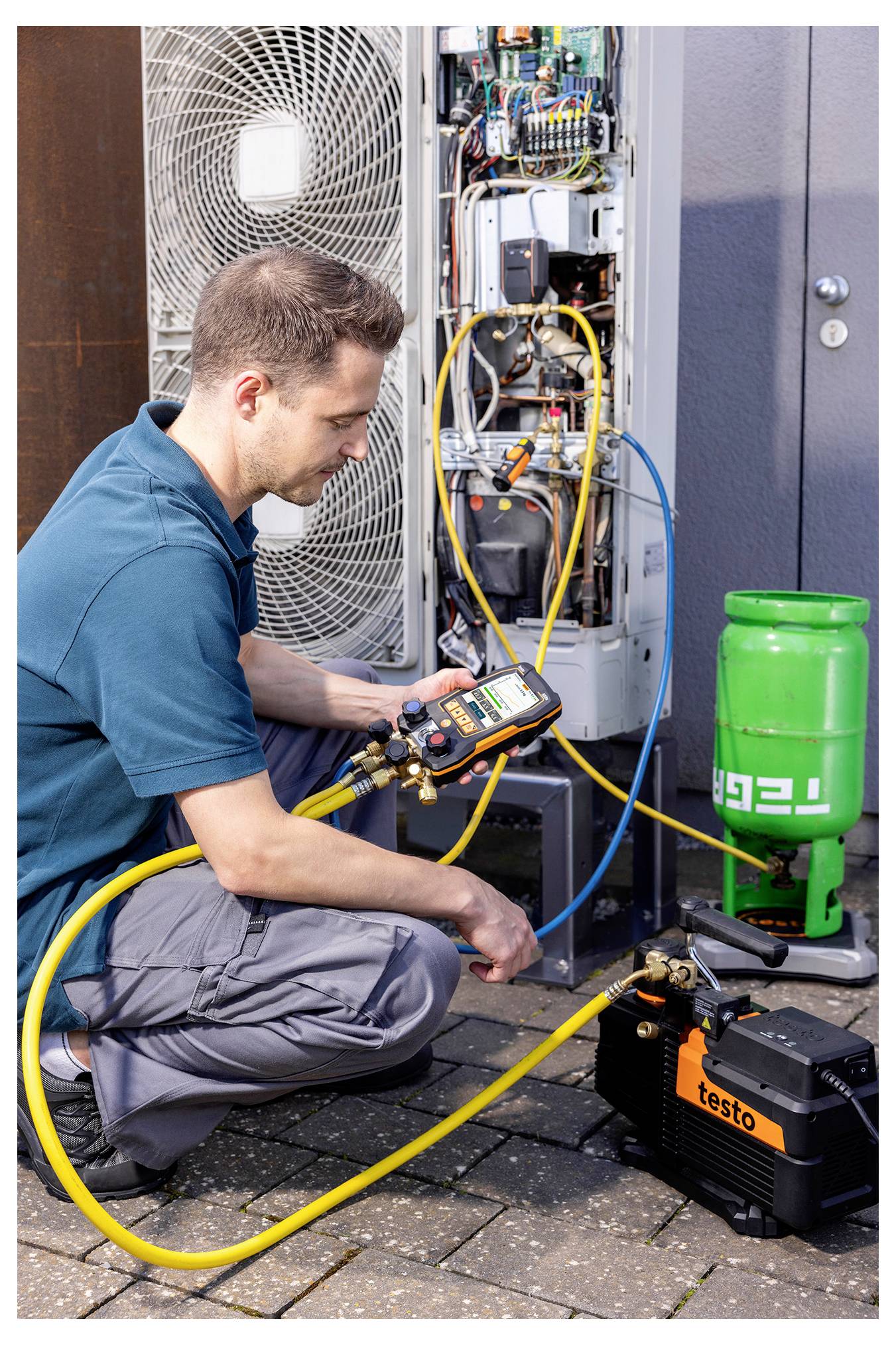 Een monteur knielt, gebruikt een digitaal manifold om een klimaatsysteem te controleren, met een koelgascilinder en compressor op de grond.