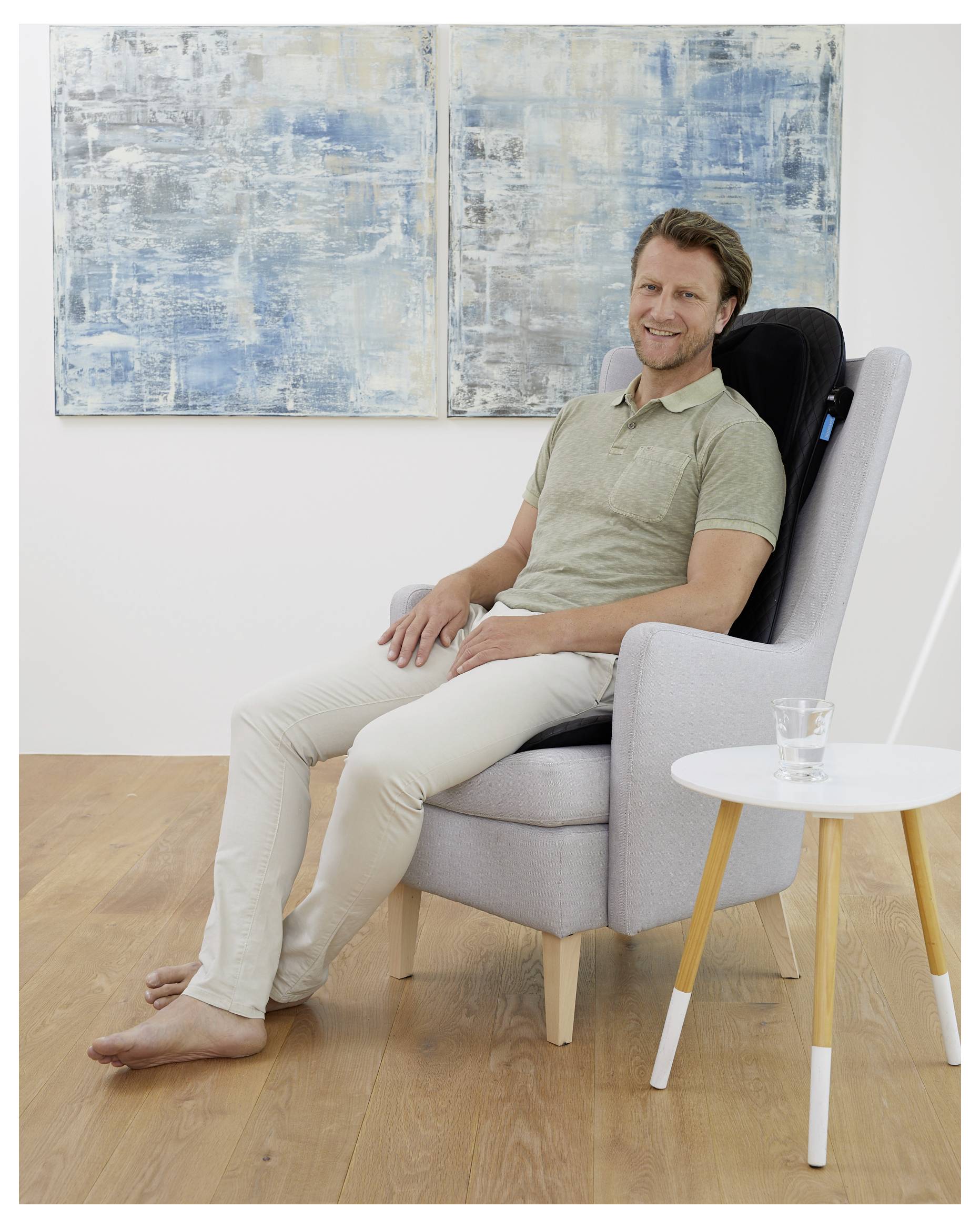 Een persoon zit blootsvoets in een lichtgrijze fauteuil, glimlachend, met een massagekussen op de rugleuning. Op een klein tafeltje staat een glas water.