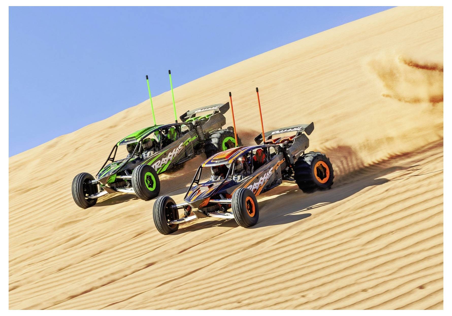 Twee terreinbuggy's met levendige ontwerpen racen zij aan zij op een steile zandduin onder een heldere blauwe lucht, waarbij ze zandsporen achter zich laten.