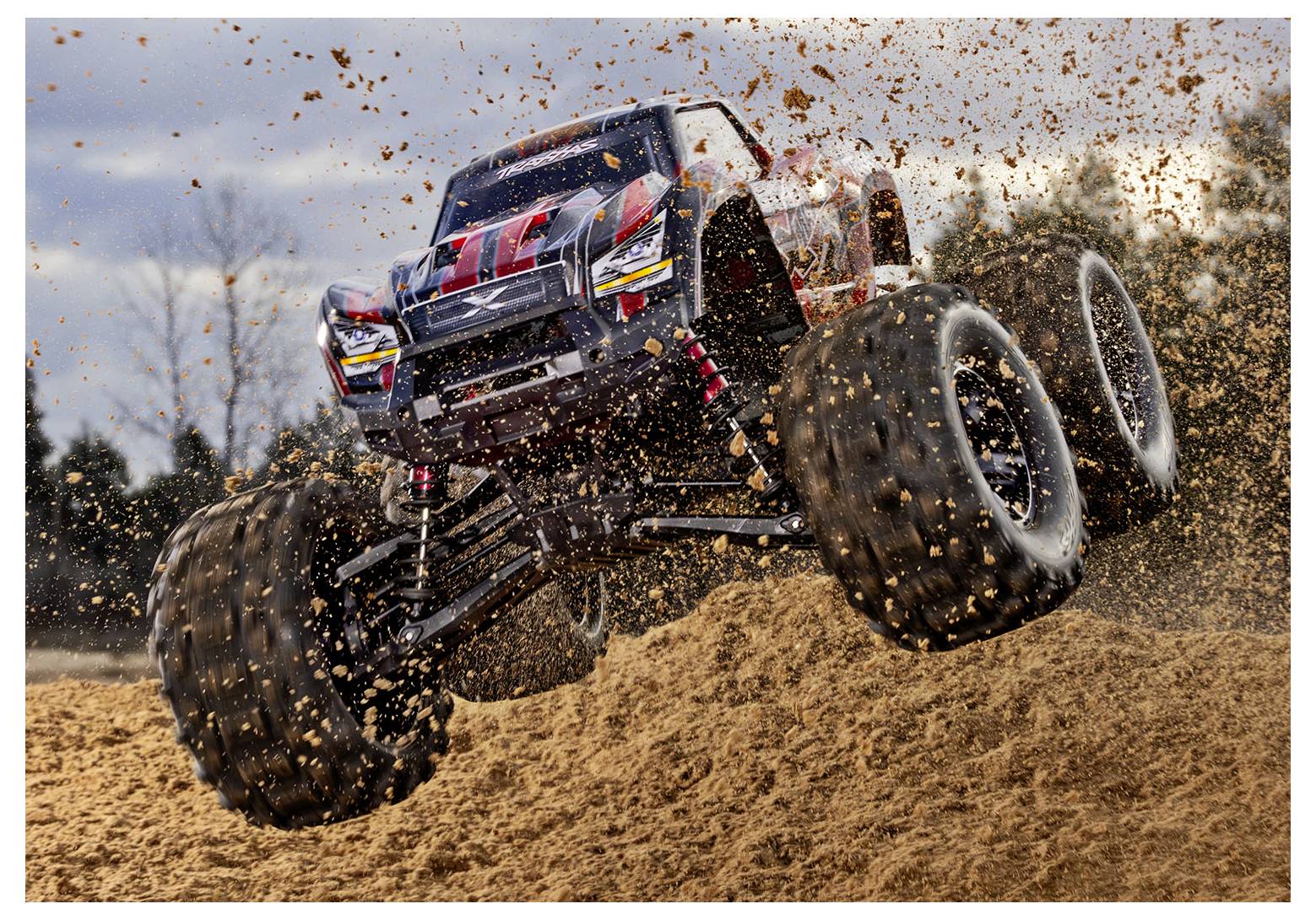 Een op afstand bestuurbare monstertruck springt over een aarden heuvel, waarbij modder wegvliegt van zijn grote banden. Bomen en een bewolkte lucht bevinden zich op de achtergrond.