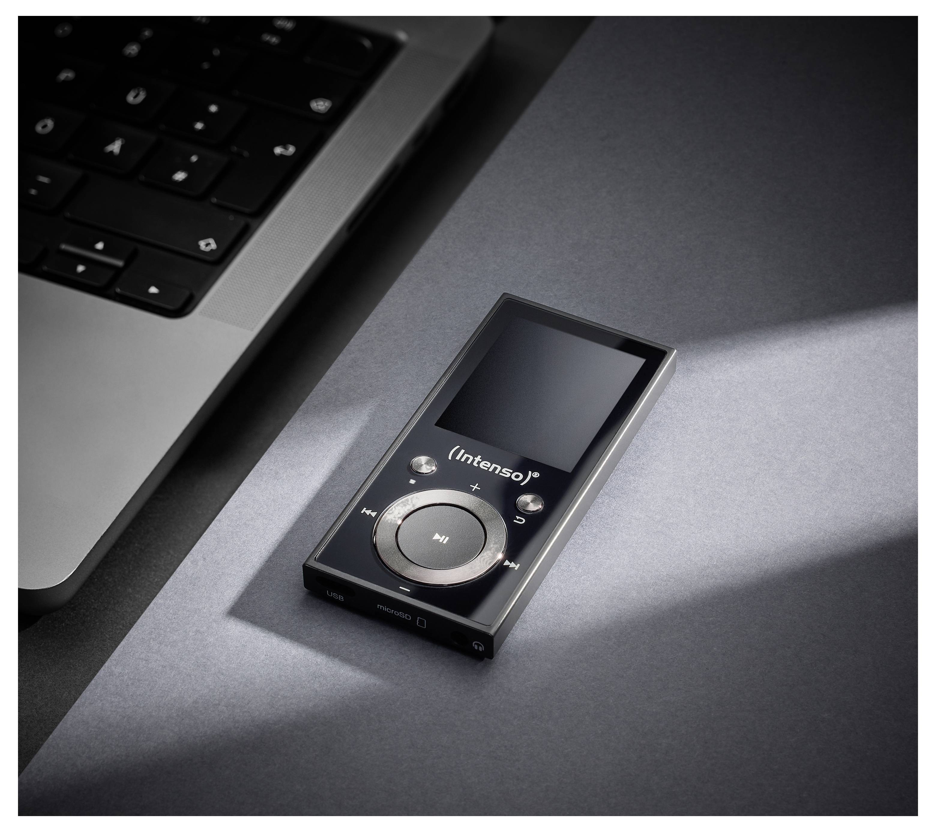Een zwarte Intenso MP3-speler met een rond bedieningsvlak rust op een grijze ondergrond nabij een laptoptoetsenbord, verlicht door een lichtstraal.