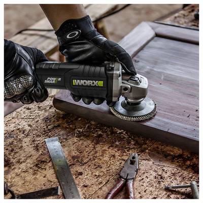 'Een persoon met handschoenen aan gebruikt een Worx elektrisch gereedschap om de rand van een houten frame op een werkbank vol gereedschap en houtsnippers te schuren.'
