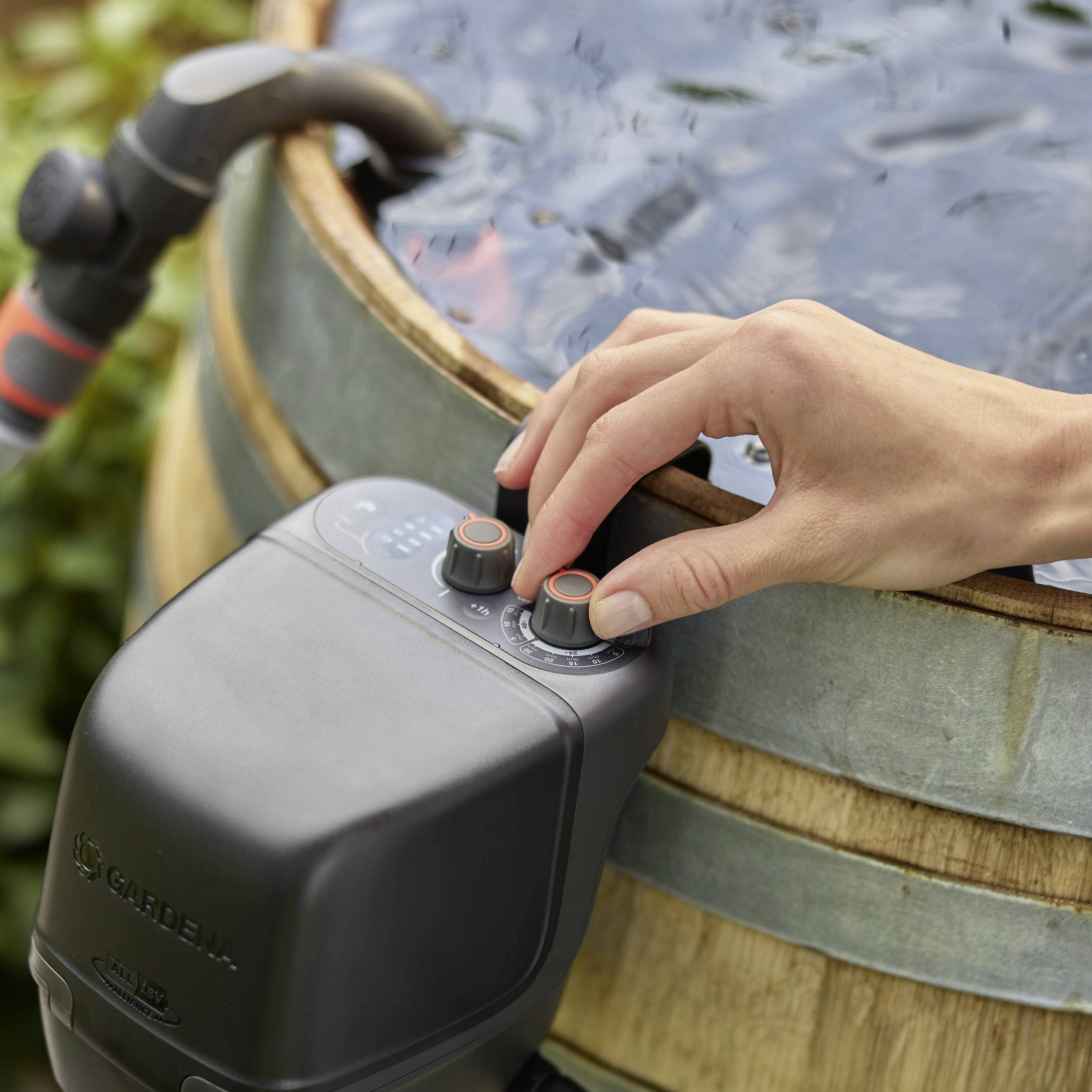 Een persoon past knoppen aan op een waterreguleertoestel naast een regenton, wat wijst op instellingen voor waterstroming of waterdruk.