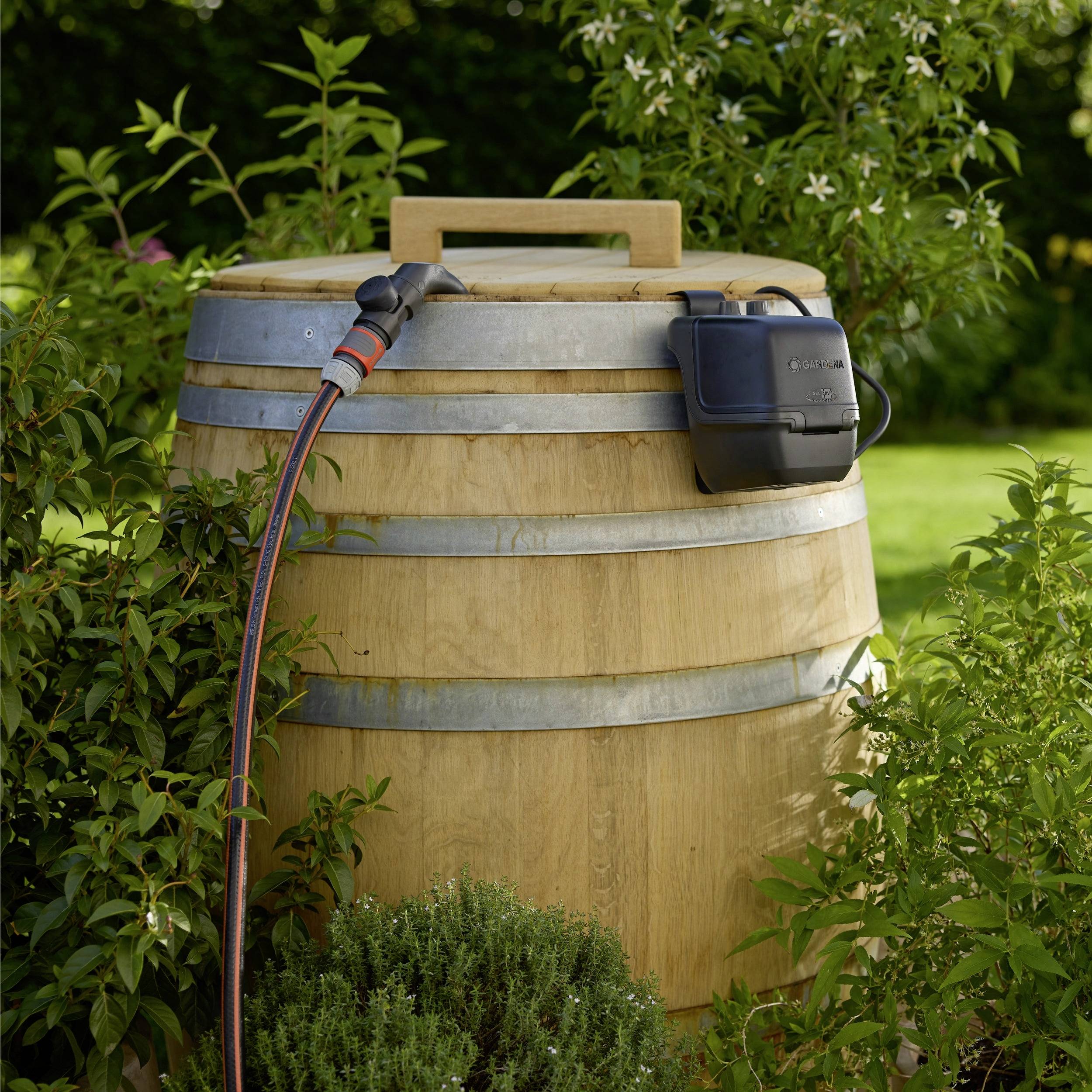 Een houten regenton met een slang eraan bevestigd staat in een tuin, omringd door groen, wat een methode voor wateropvang demonstreert.