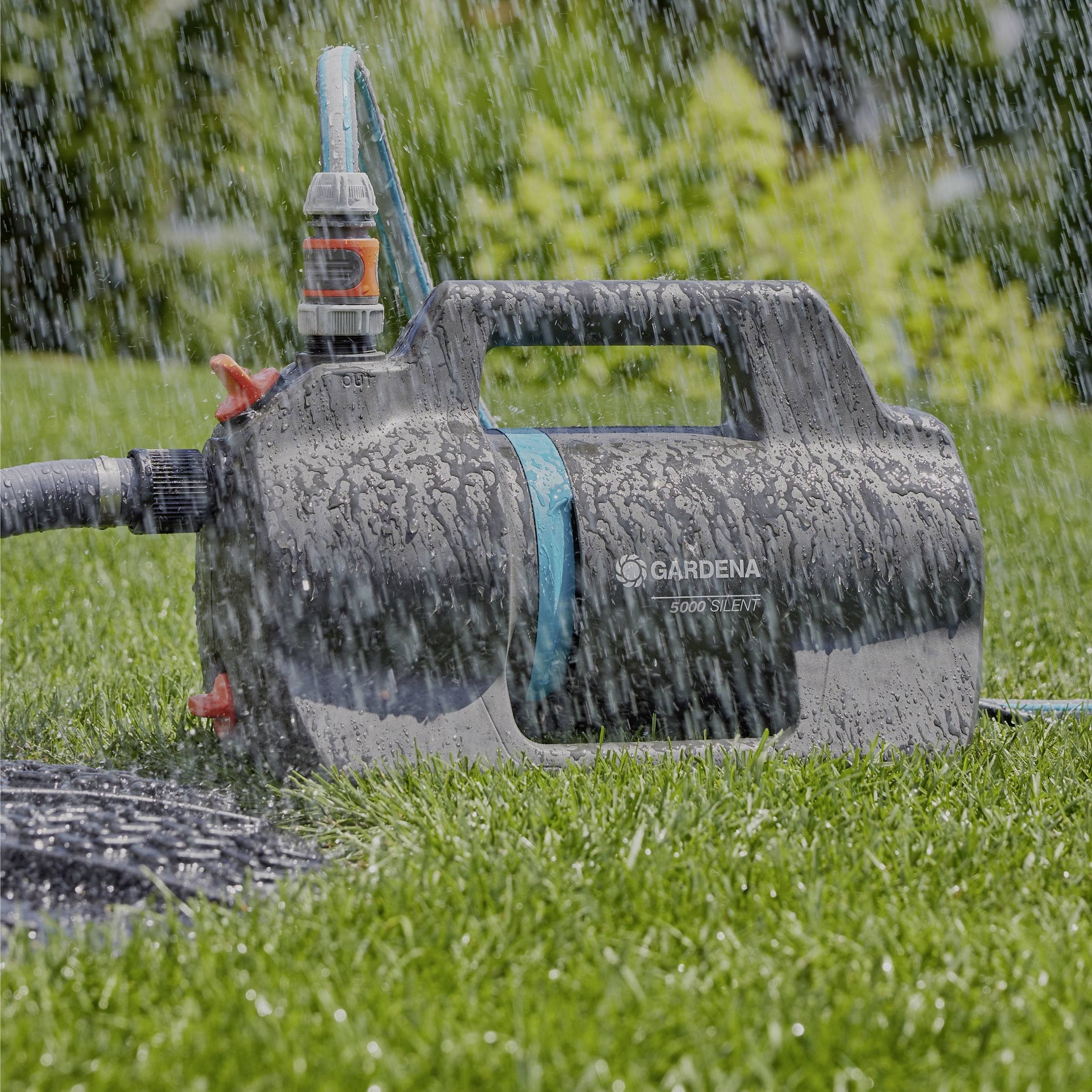 Een draagbare, natte waterpomp op gras, gelabeld 'Gardena', met een slang aangesloten, gebruikt voor tuinirrigatie in een regenachtige buitenomgeving.