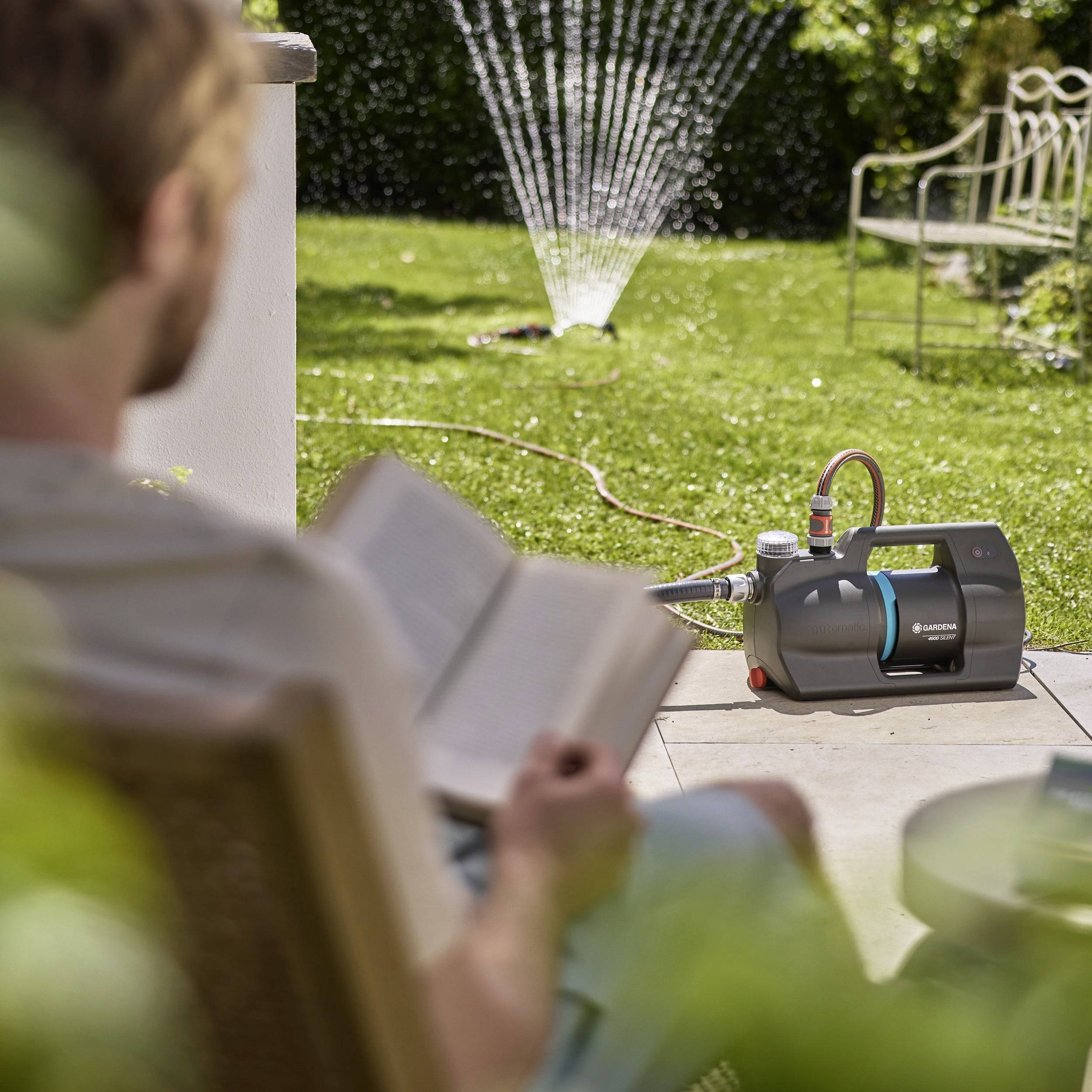 Een persoon die een boek leest op een patio met een tuinfontein die op de achtergrond loopt. Een waterpomp is zichtbaar op het gras in de buurt.